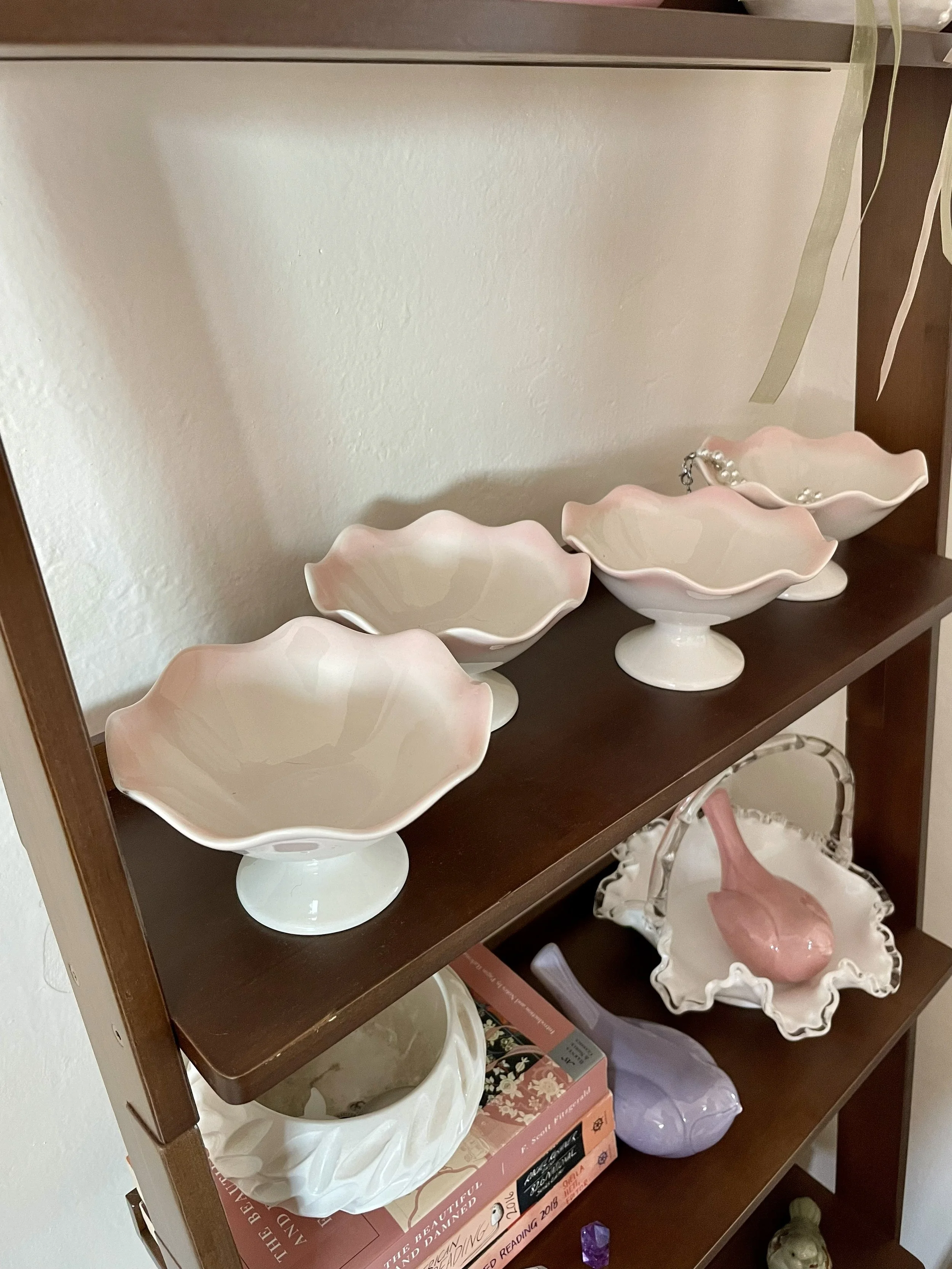 A wooden shelf with four pastel-colored ceramic bowls. A decorative pink ceramic bird in a white decorative basket, a white ceramic bowl, a purple ceramic bird, and a stack of pink books on the lower shelf.