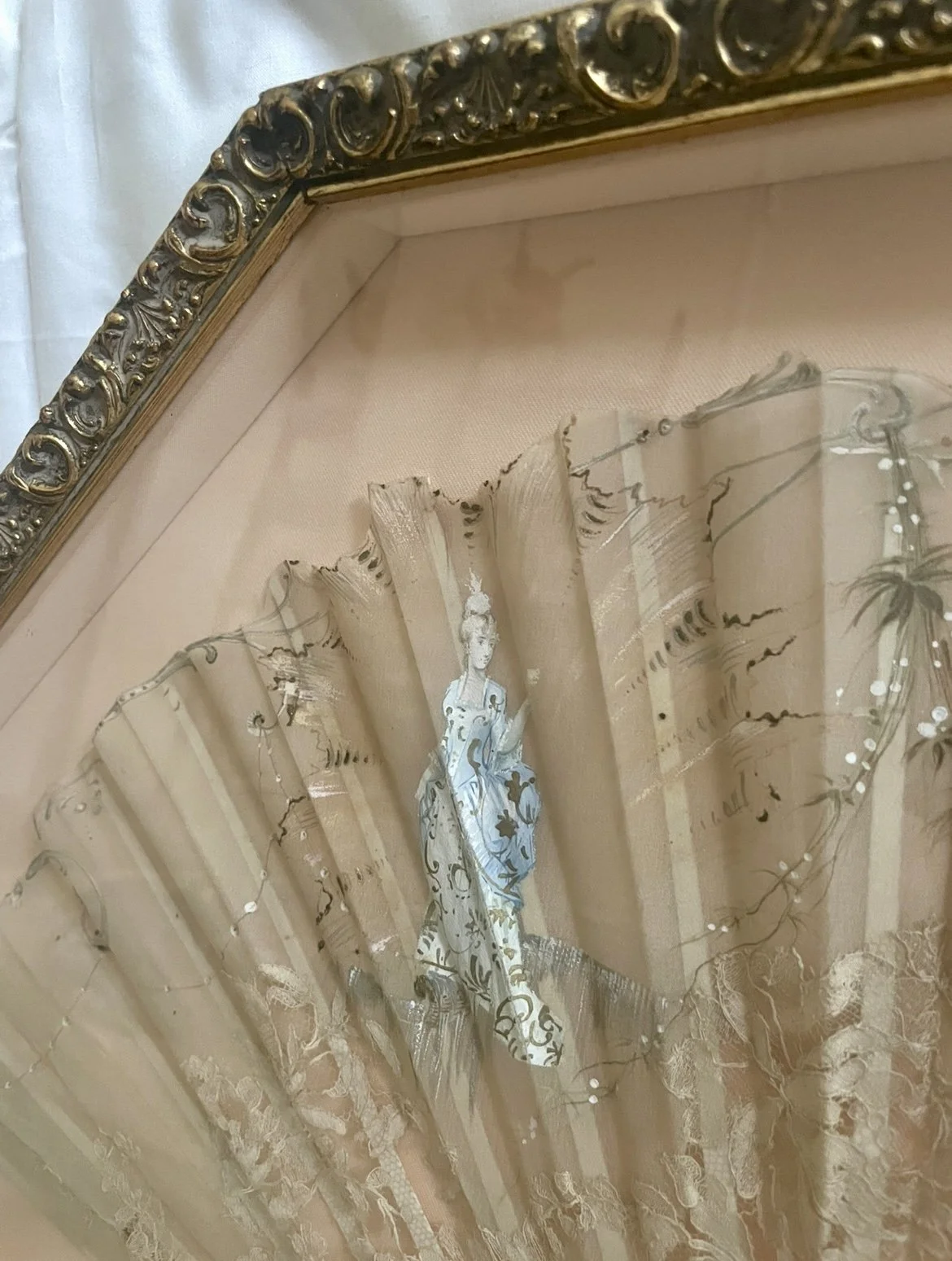 A decorative box with an ornate gold detailed trim, containing a delicate, antique hand fan with a painted woman on its fabric. The hand fan fabric is light-colored with floral and artistic designs.