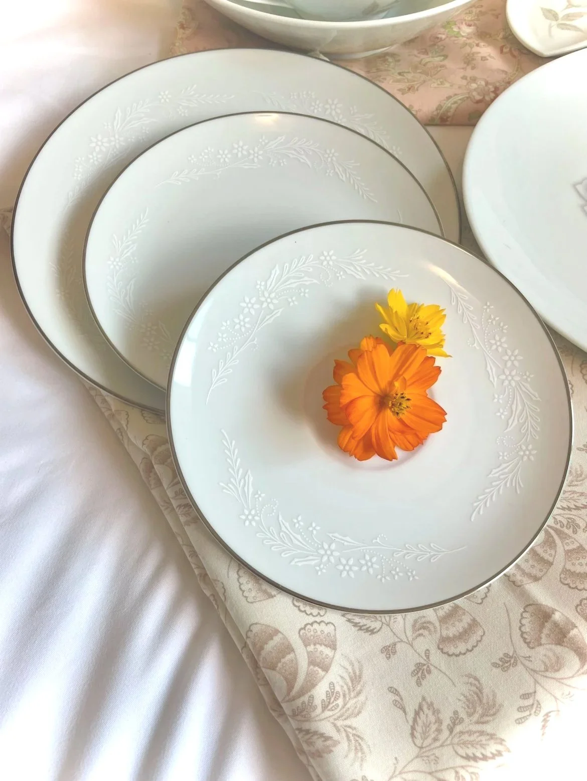 White porcelain plates with silver rims and floral patterns, arranged on a table with a pink patterned tablecloth. One plate holds two orange and yellow flowers.