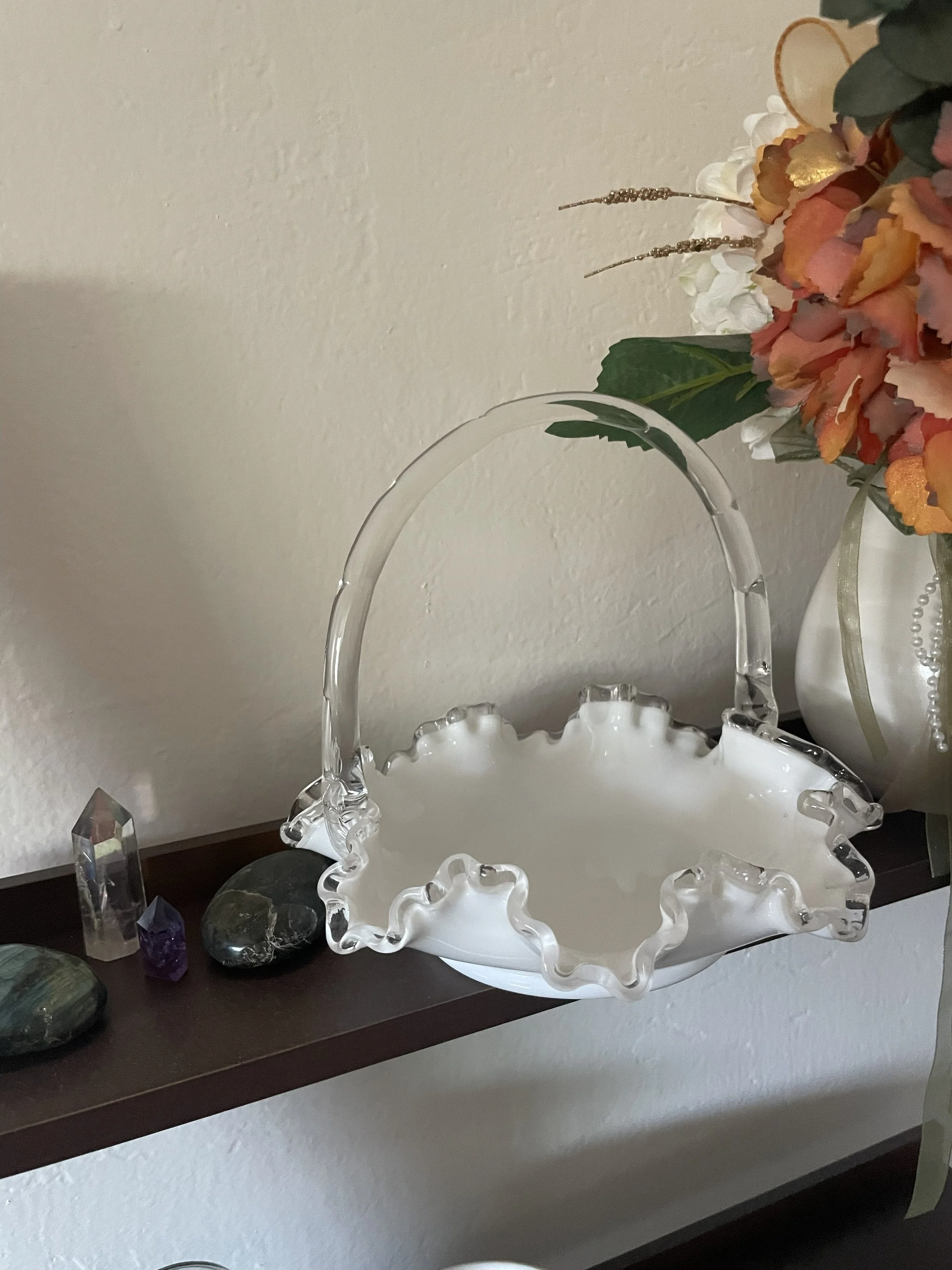 A decorative white glass basket with ruffled edges and a clear handle, sitting on a dark wooden shelf next to a bouquet of flowers in shades of orange, pink, and white.