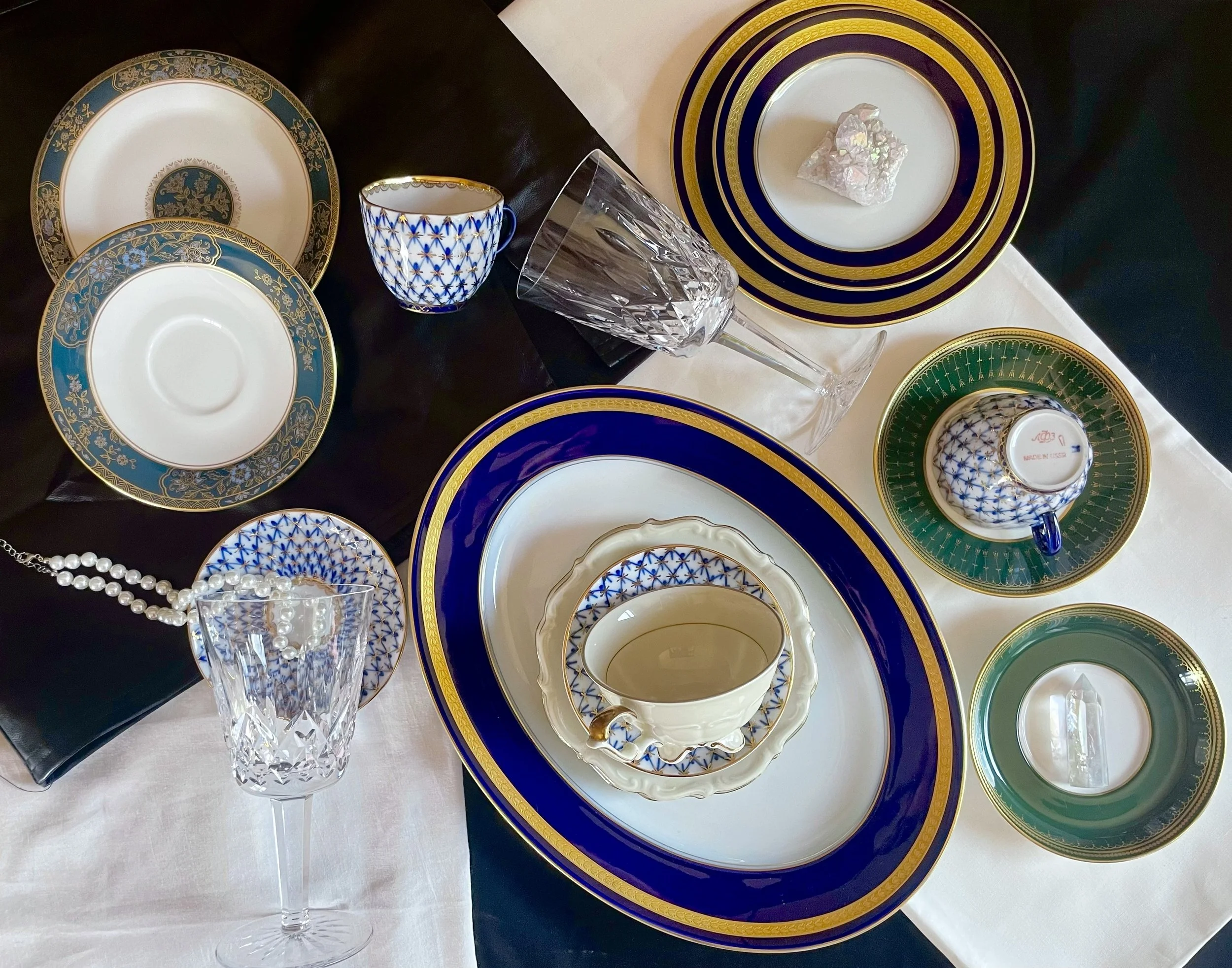 Mismatched vintage china united by repeating gold rims and a recurring blue net motif, with gold-rimmed plates, blue-and-gold cups and saucers, and cut crystal on white and black linen.