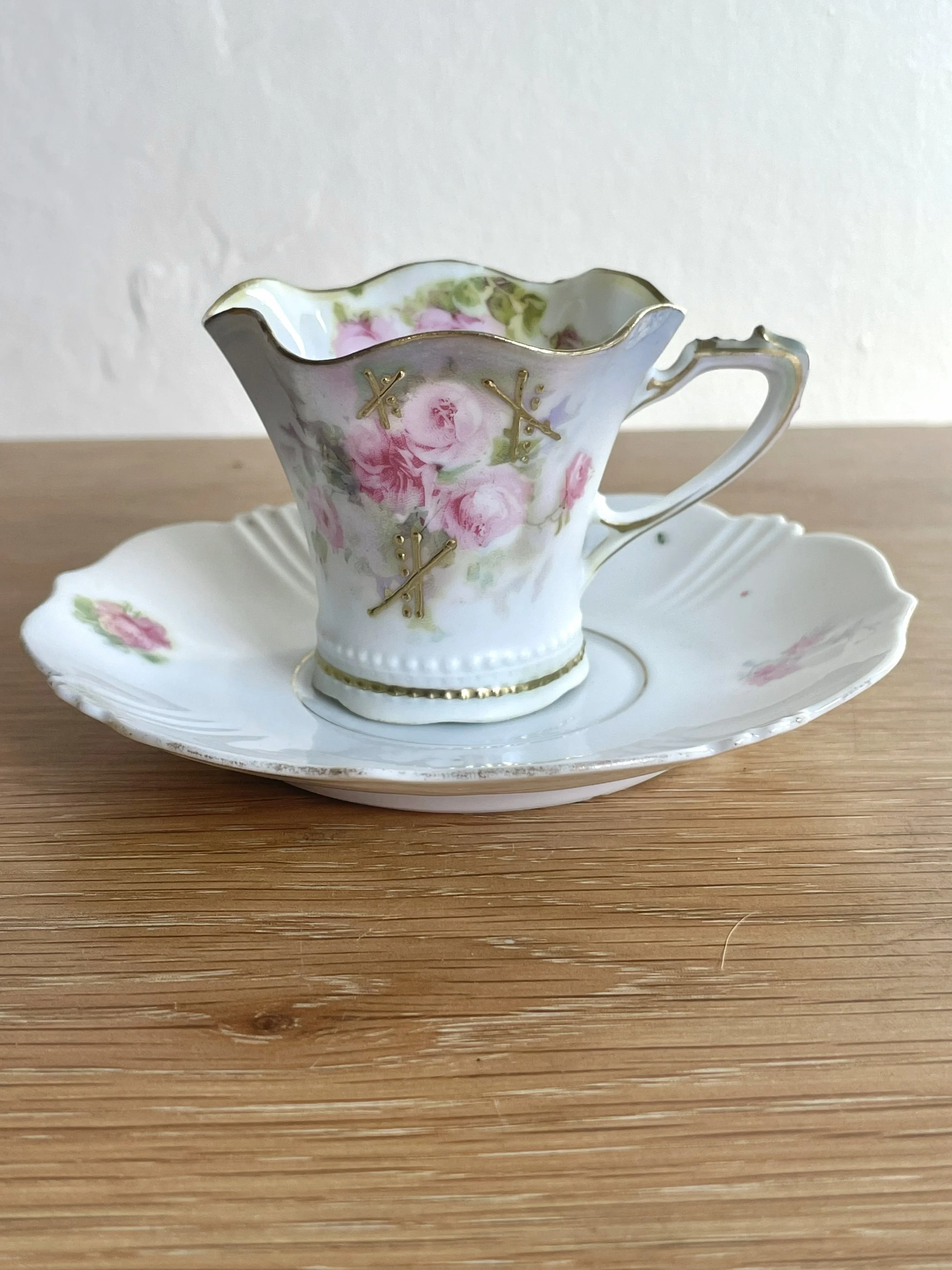 A vintage porcelain teacup with pink roses and gold accents, sitting on a matching saucer with pink roses, placed on a wooden surface.