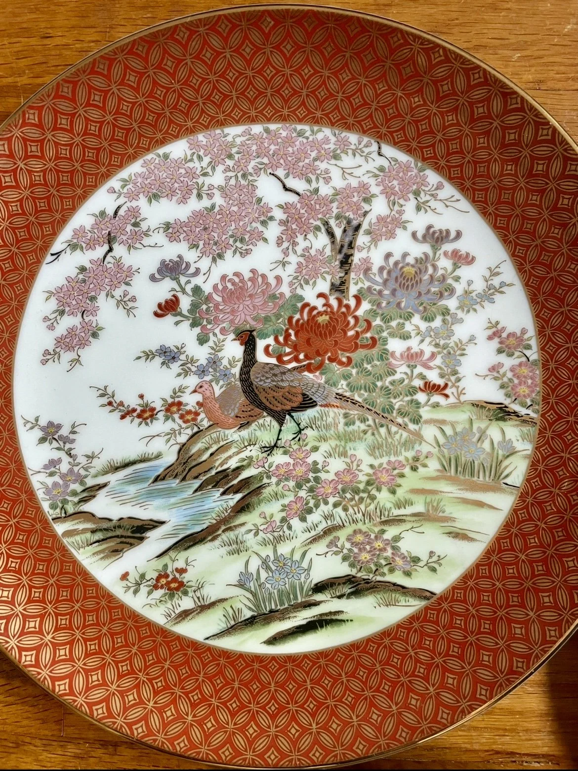 Decorative porcelain plate with a painted scene of pheasants by a stream, surrounded by pink cherry blossoms and other flowers, with a red and gold patterned border.