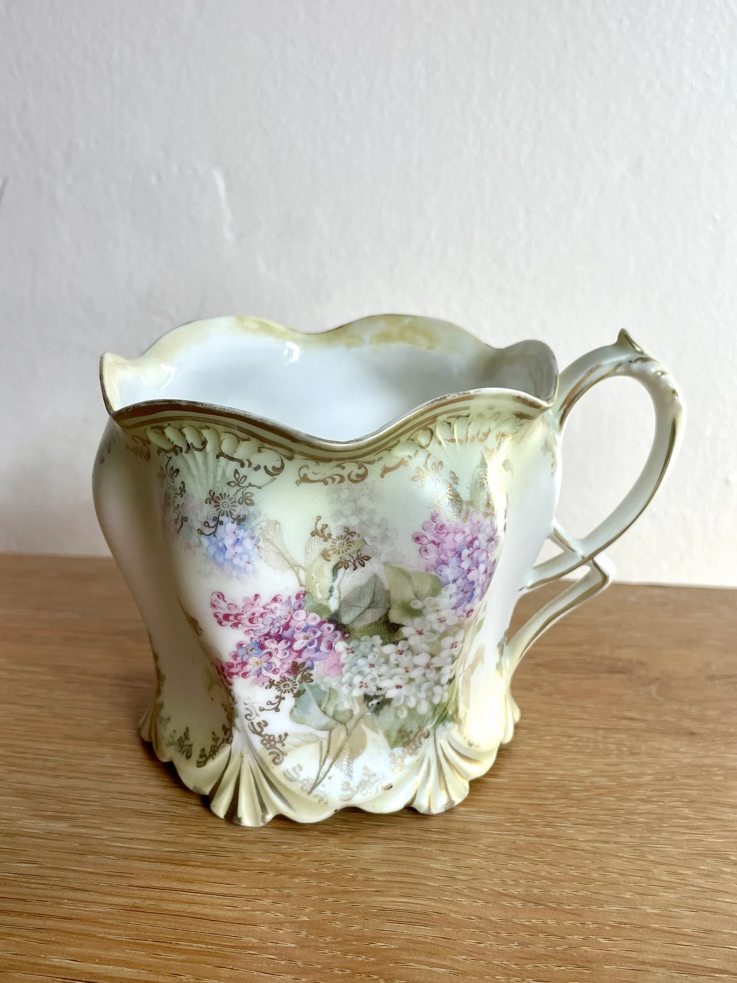 A vintage floral ceramic pitcher with a decorative handle, on a wooden surface against a plain wall background.