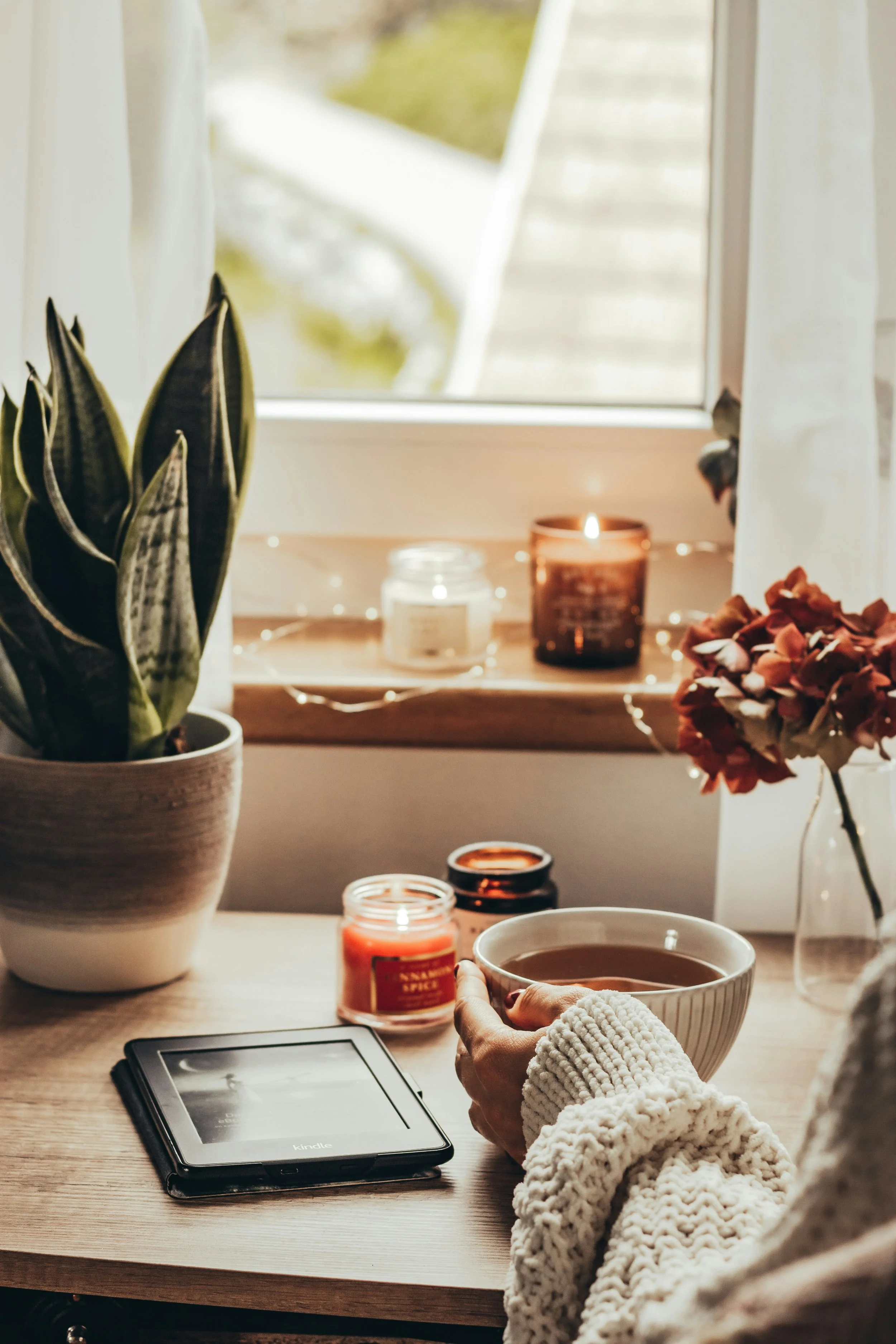 A hand holds a large cup of tea on a table with candles, a potted plant, dried flowers, and an e-reader, creating a cozy indoor scene.
