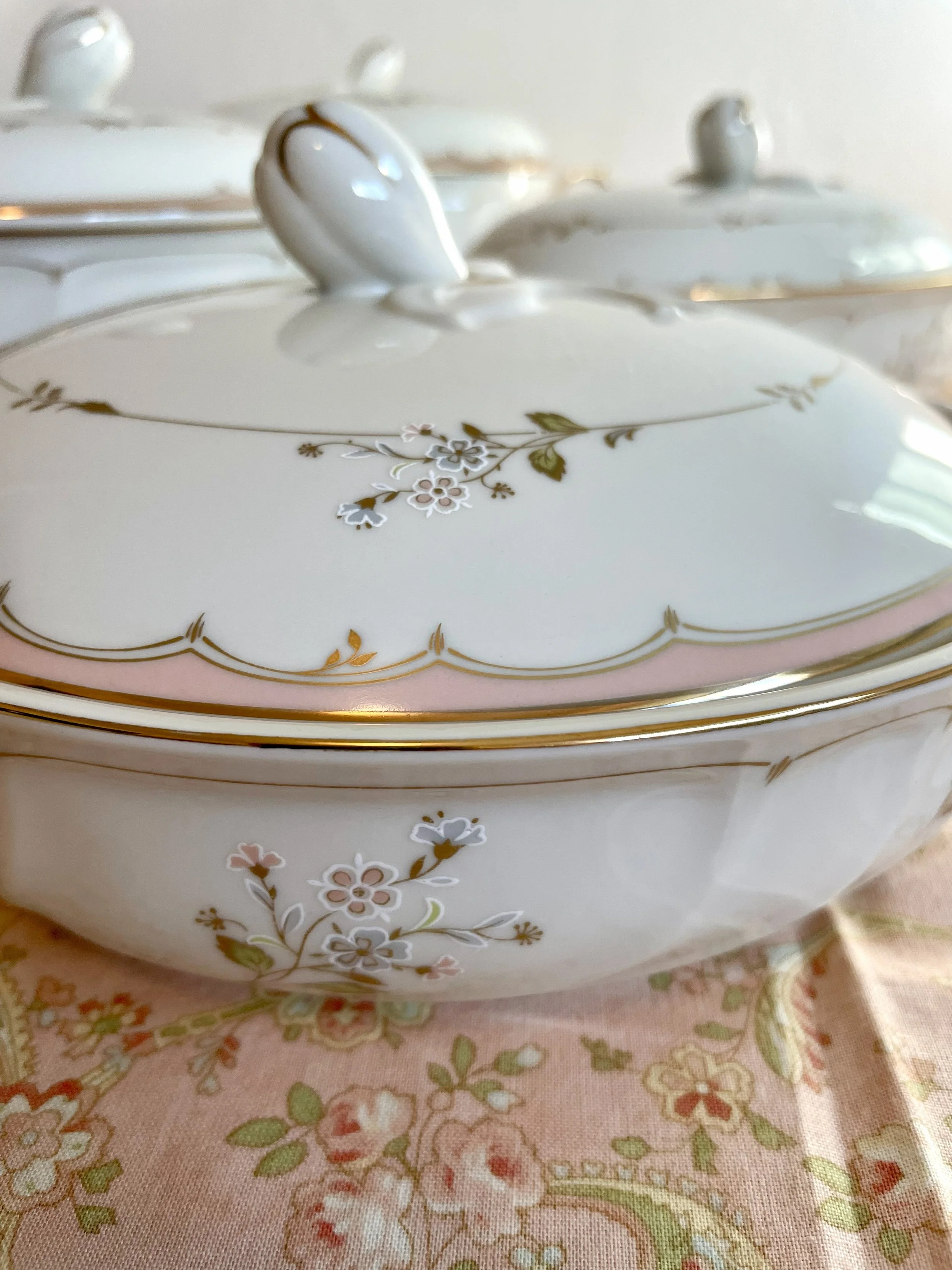 Close-up of a white porcelain covered serving dish with a floral design and gold trim, placed on a pink floral tablecloth.