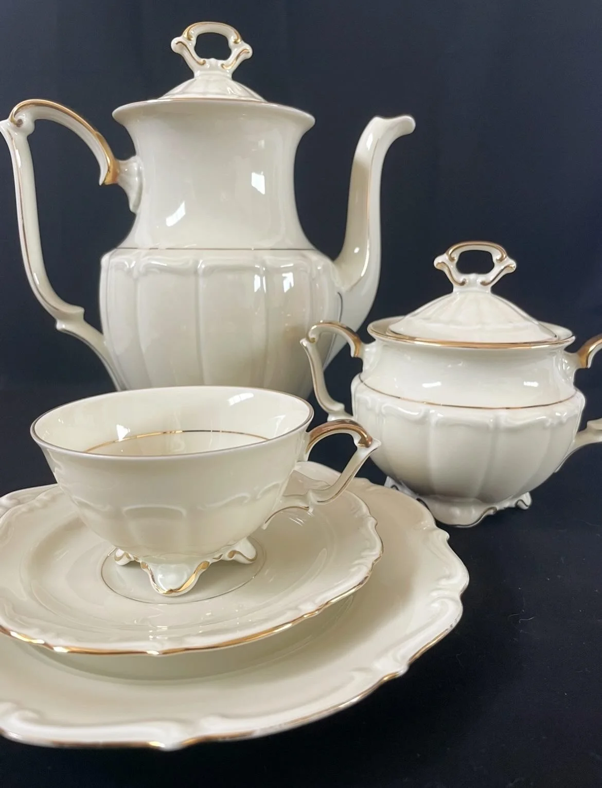Elegant porcelain tea set with gold trim, including a teapot, sugar bowl with lid, teacup and saucer, displayed against a dark background.