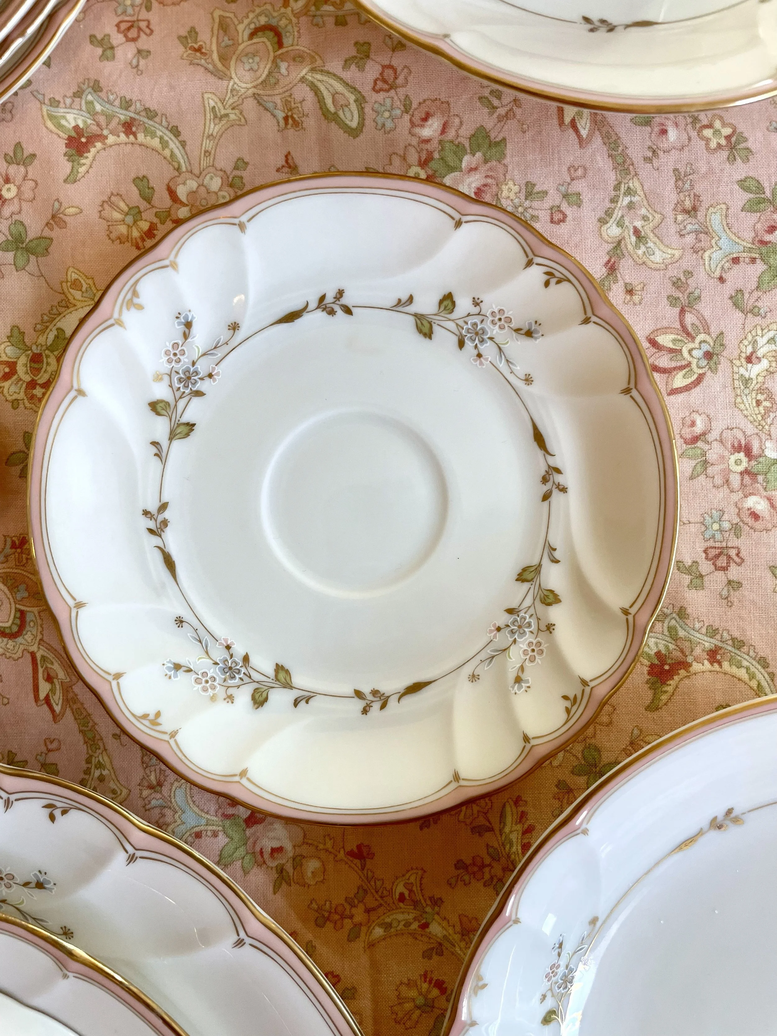 Elegant ceramic plate with a scalloped edge, pink and gold trim, and a floral pattern of small white and pink flowers with green leaves, placed on a pink floral tablecloth.
