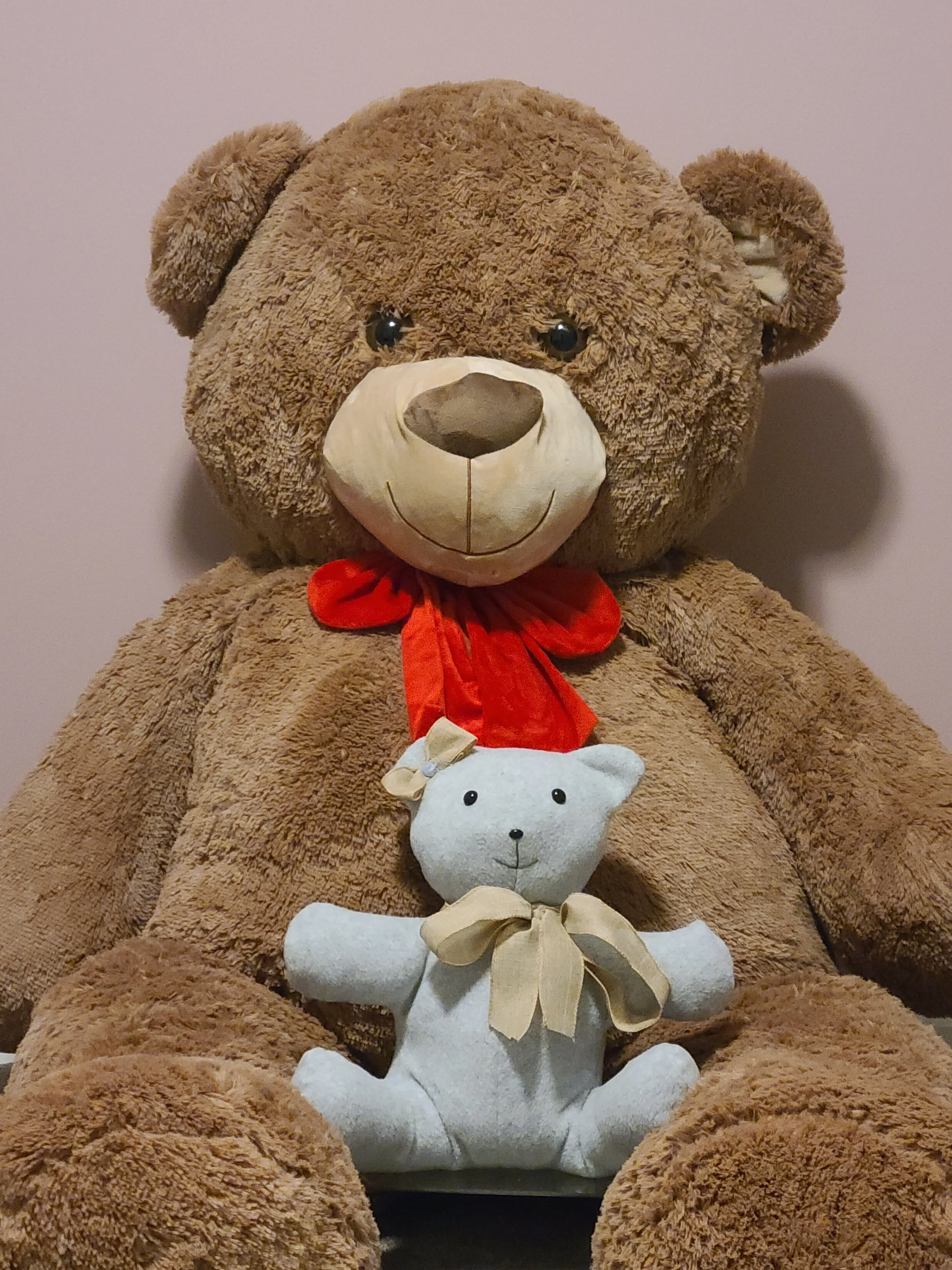 A large plush teddy bear with a red bow tie and a smaller white teddy bear with a beige bow tie sitting in front of it.