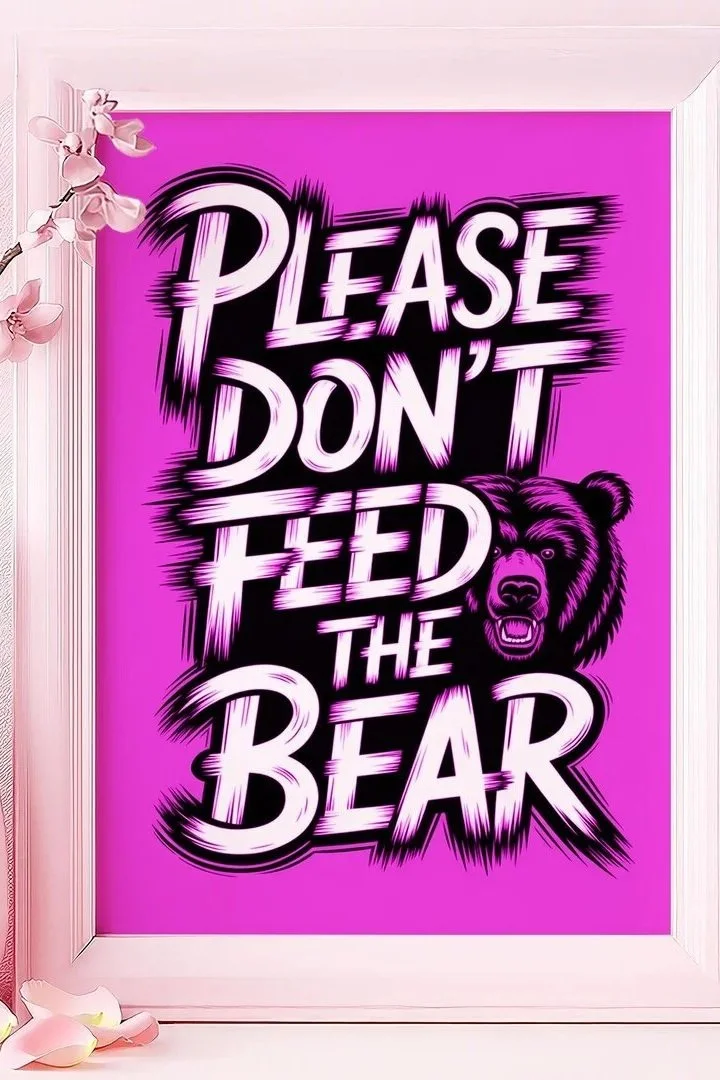 Pink sign with bold white and black text reading "Please Don't Feed the Bear" and an illustration of a bear's head, framed by white borders and decorated with pink flowers.