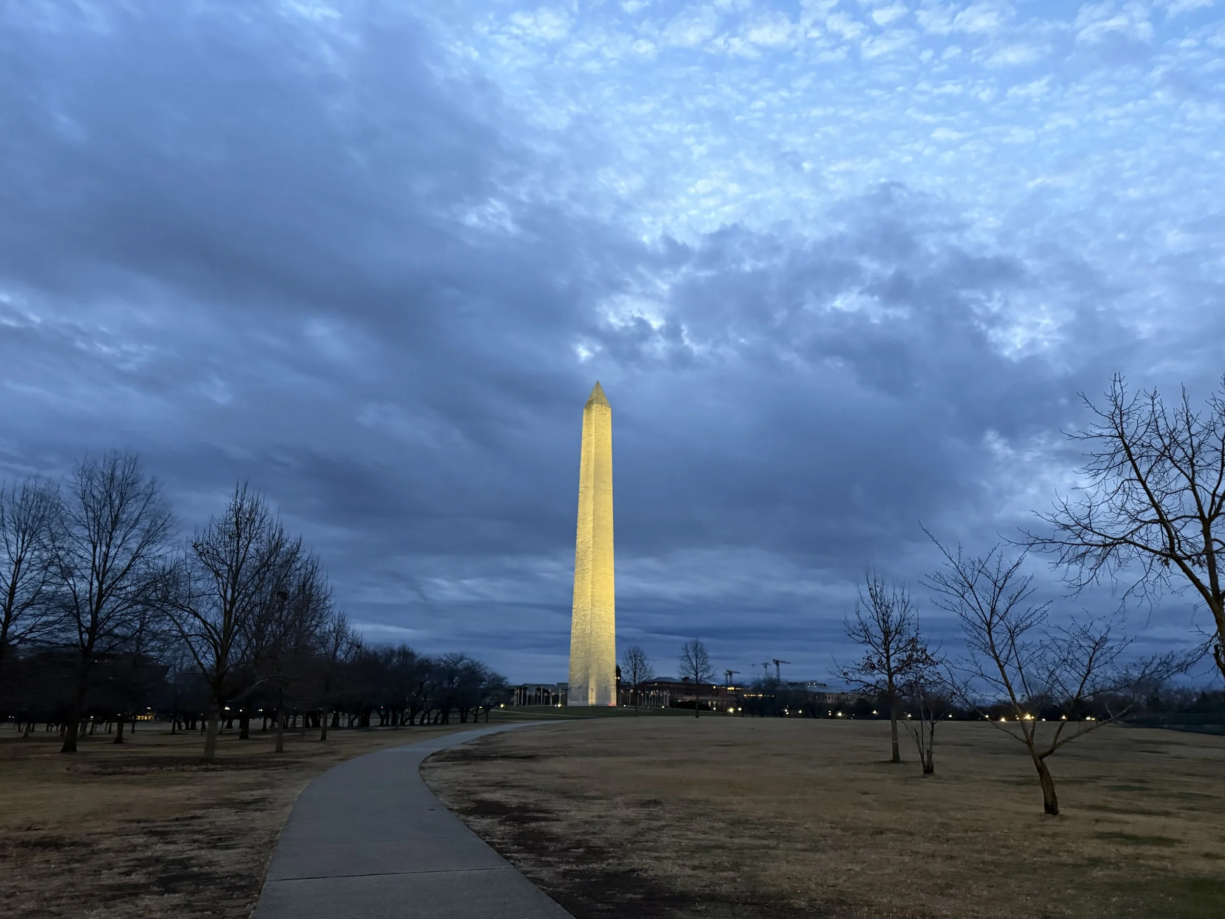 Washington Monument lit up at dusk, with a cloudy sky overhead and leafless trees surrounding a pathway in a park.