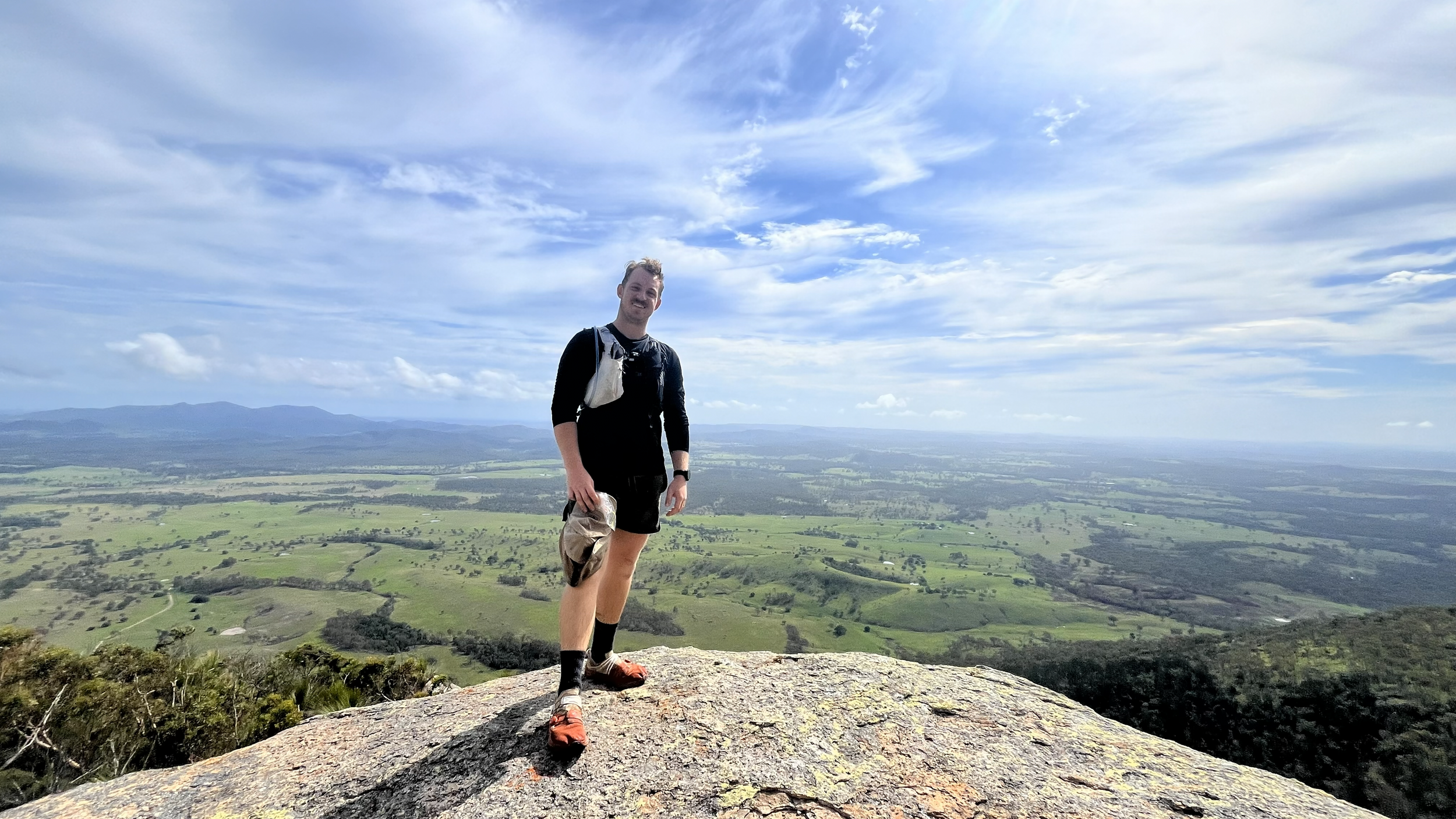 A man standing on a large rock at the top of a mountain, overlooking a vast landscape of green fields and distant mountains under a partly cloudy sky.