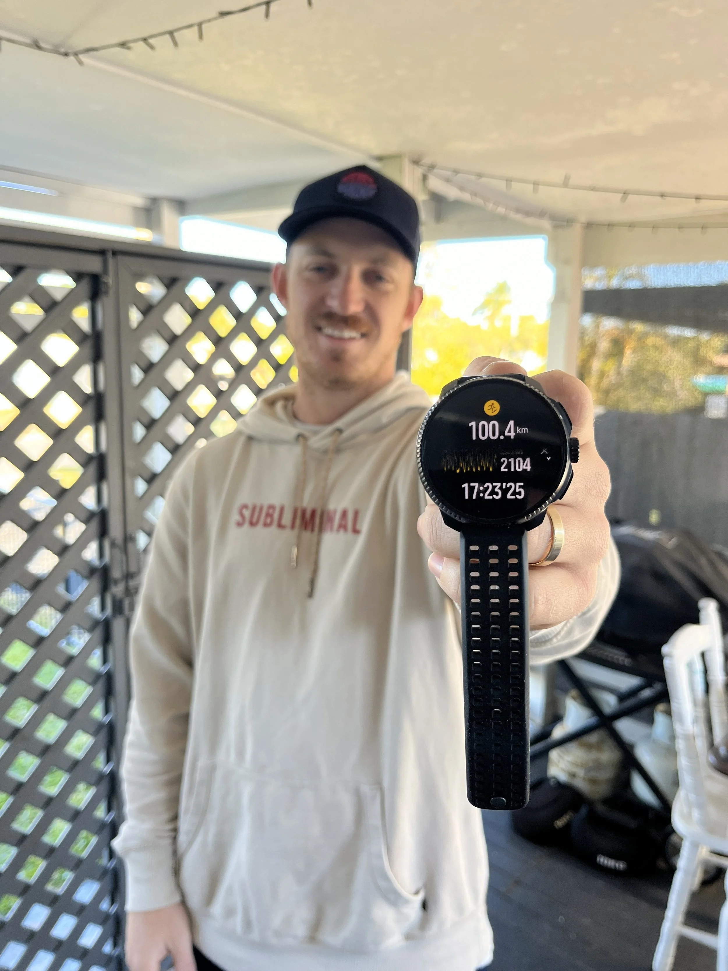 A man is smiling and holding up a smartwatch displaying 100.4 km, 2104 calories, and 17:23:25. He is wearing a black cap and a beige hoodie with 'SUBLIMINAL' written on it. The background shows an outdoor area with a lattice fence and some trees.