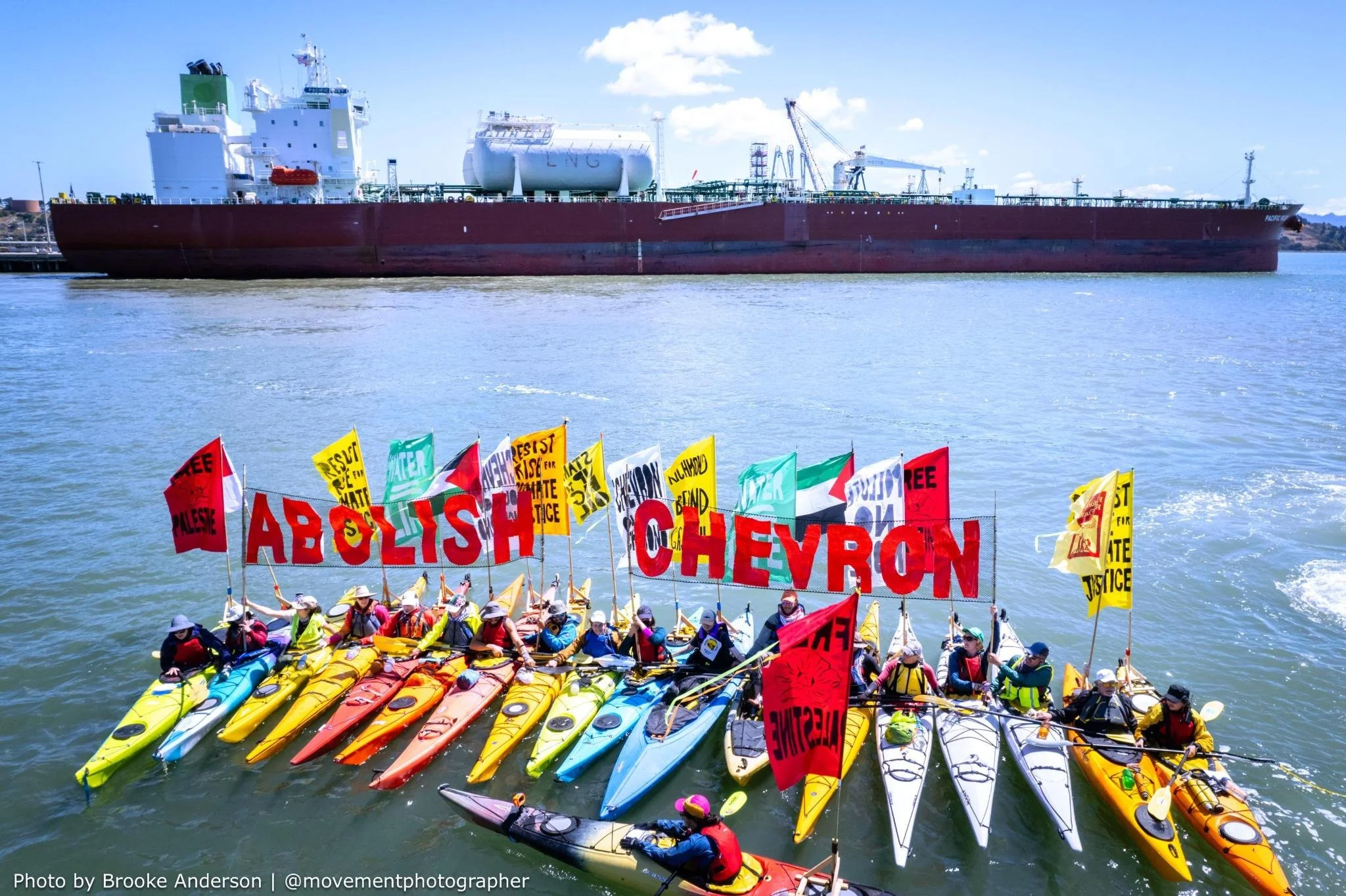 A colorful floatilla of 18 kayaks aligned side by side hold banners reading "Abolish Chevron" and many colorful flags with many messages including palestinian flags