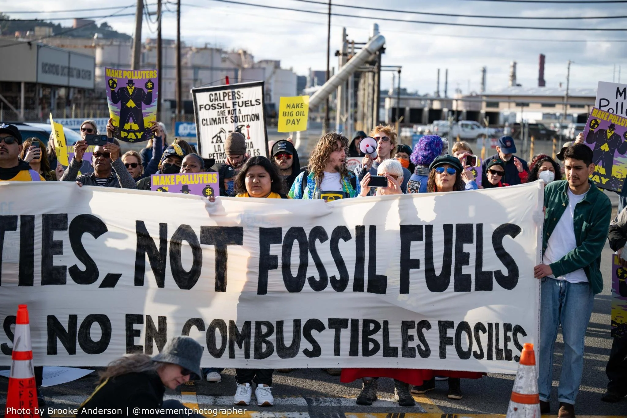 People holding signs reading make polluters pay and a banner saying Not fossil fuels in front of the Chevron refinery in Richmond