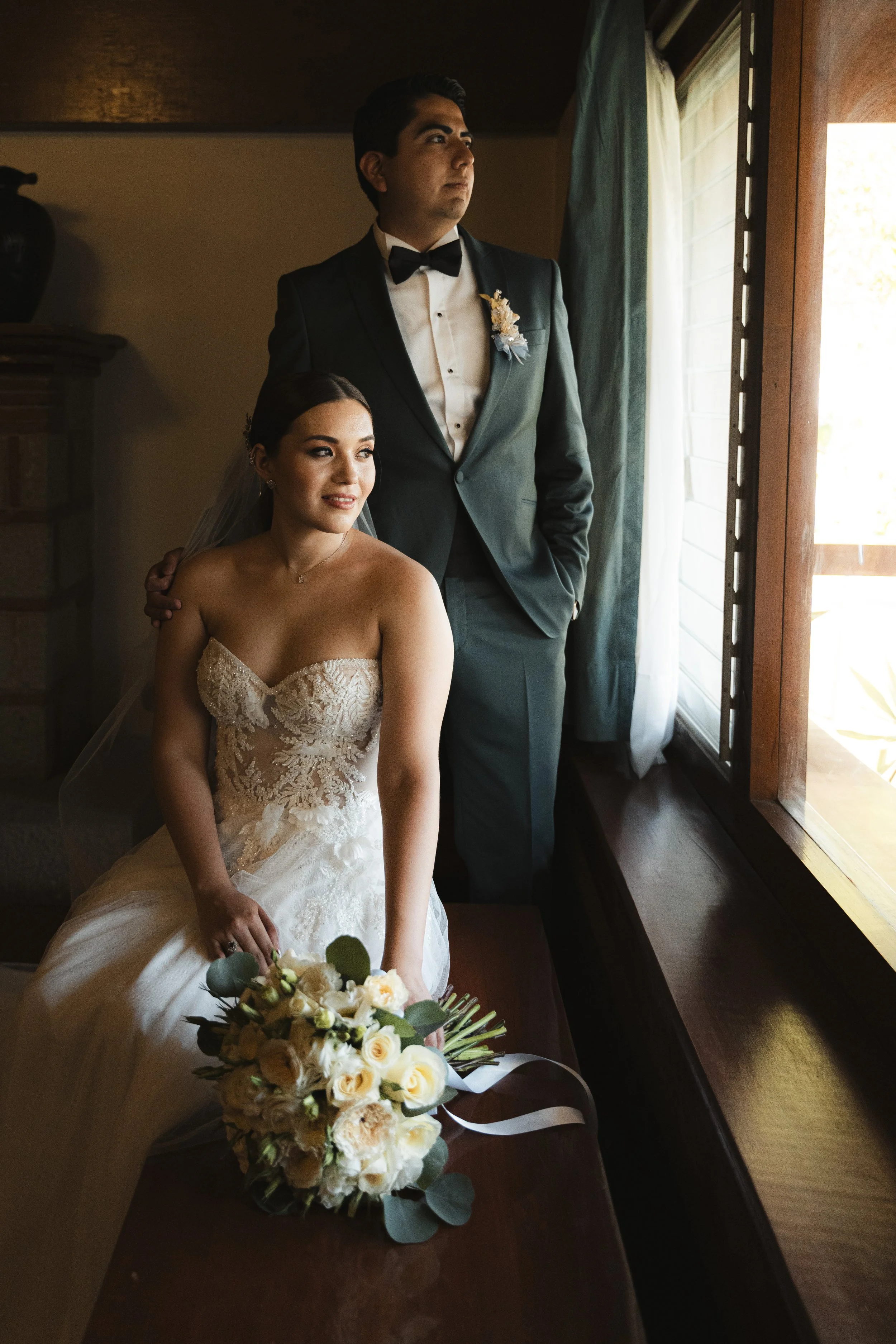 Una pareja de novios en su boda, la novia sentada con un ramo frente a ella, y el novio de pie mirando por la ventana, en un ambiente interior con luz natural