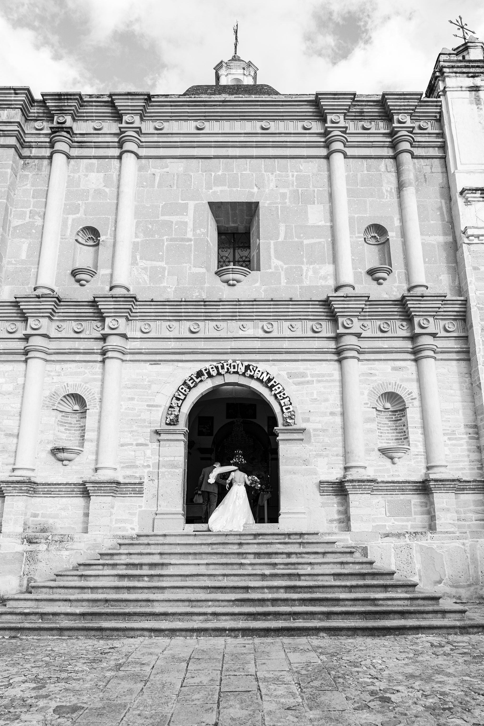 Pareja de novios en la entrada de una iglesia colonial, con escaleras y estructura de piedra, en un día nublado.
