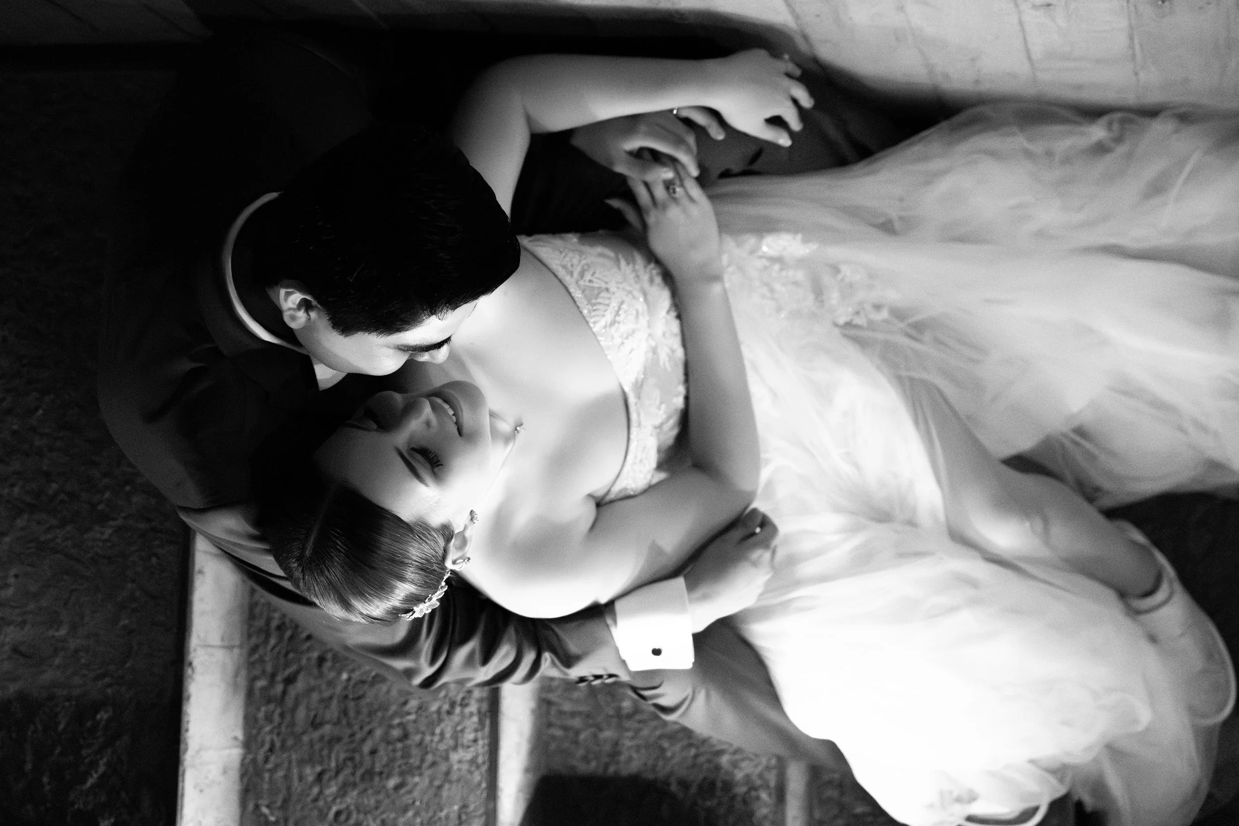 Pareja en boda, el hombre inclinado hacia la mujer acostada en el suelo, ambos sonriendo, en un entorno oscuro. La mujer lleva vestido de novia y la escena es en blanco y negro.