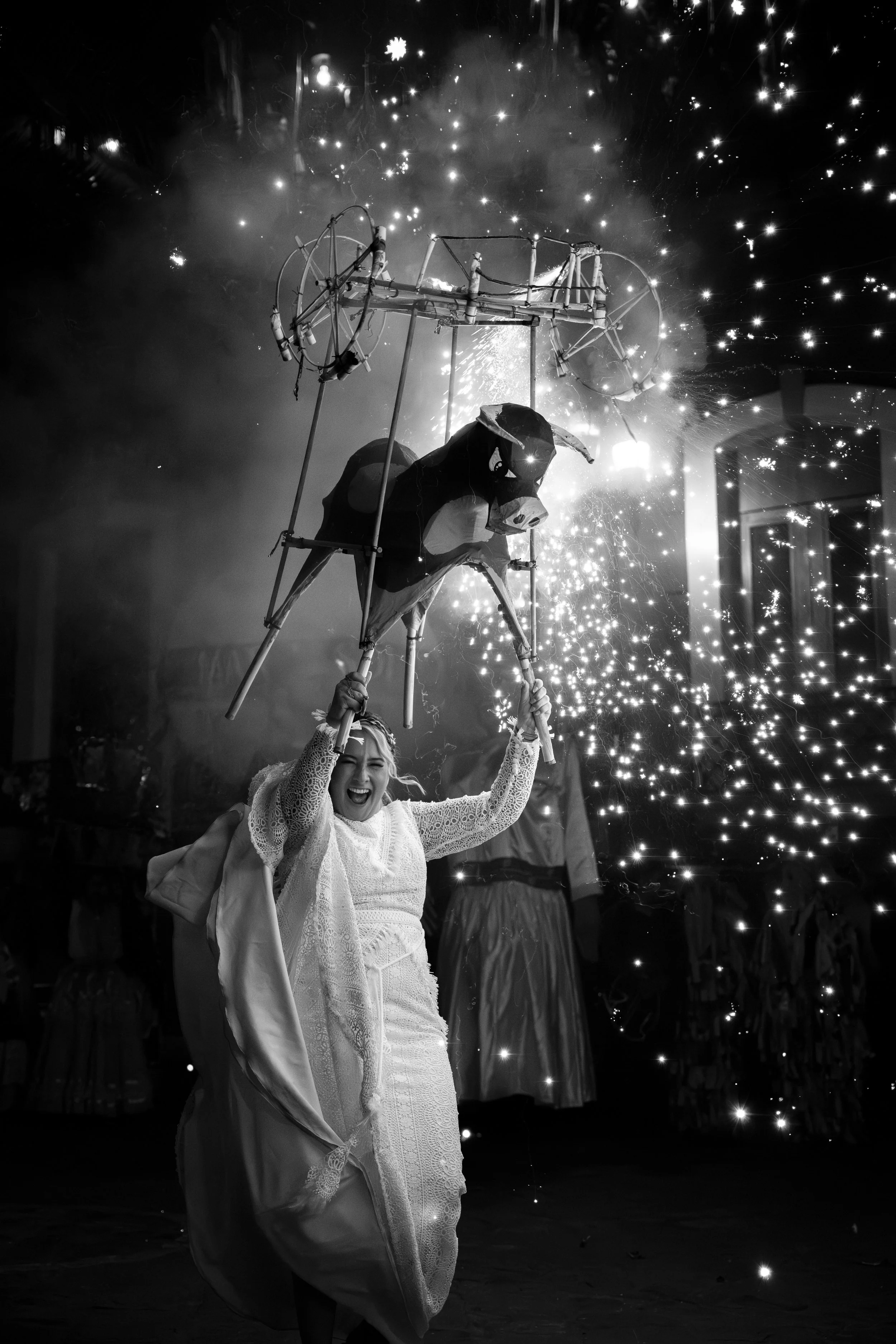 Una mujer sonriendo, sosteniendo una estructura de marioneta de un perro con fuegos artificiales de fondo en un evento festivo.