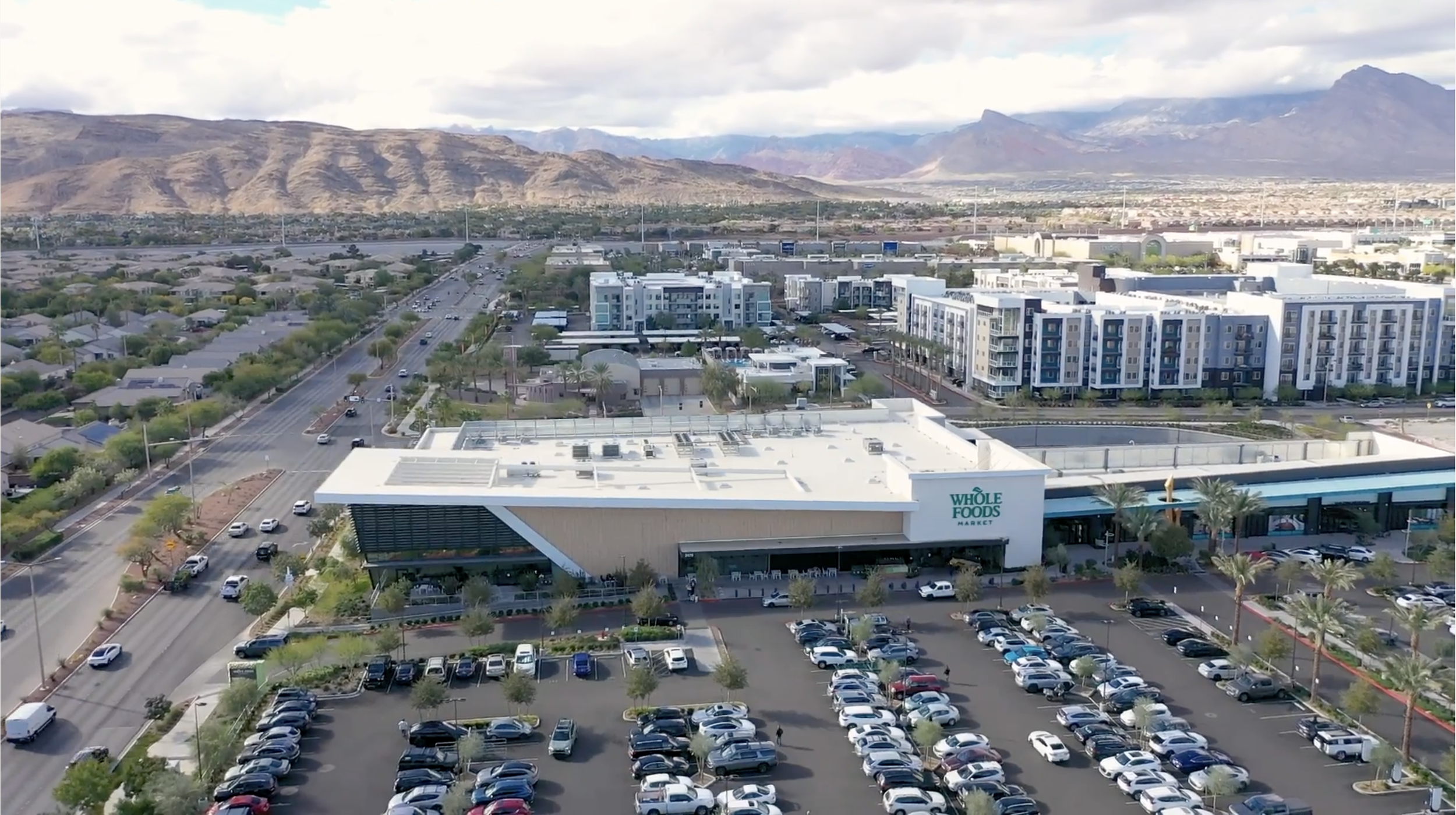 Aerial view of a Whole Foods Market parking lot with numerous cars, a shopping plaza, surrounding residential area, and mountains in the background.