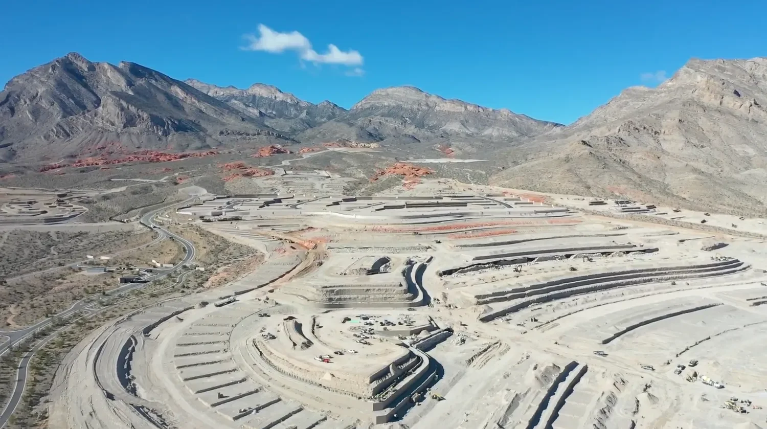 An aerial view of a mining or construction site with multiple terraced levels in a mountainous desert landscape under a blue sky.