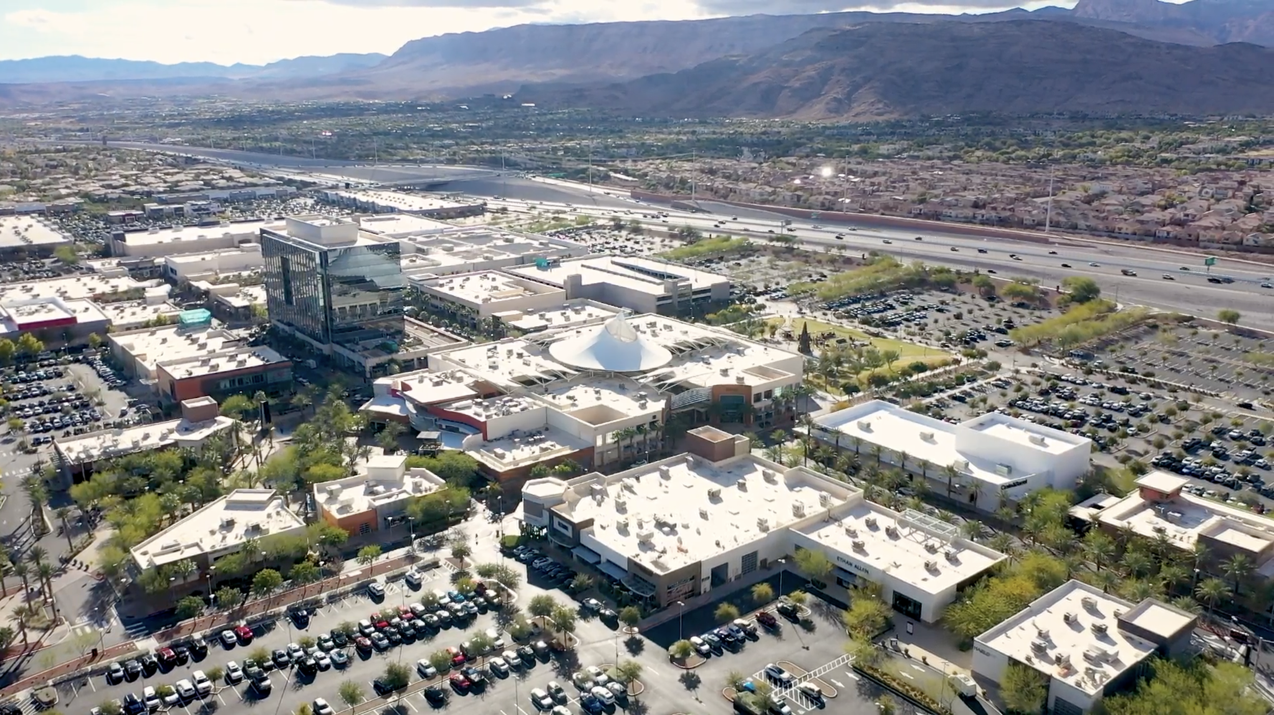 An aerial view of a cityscape with commercial buildings, parking lots filled with cars, and mountains in the background.