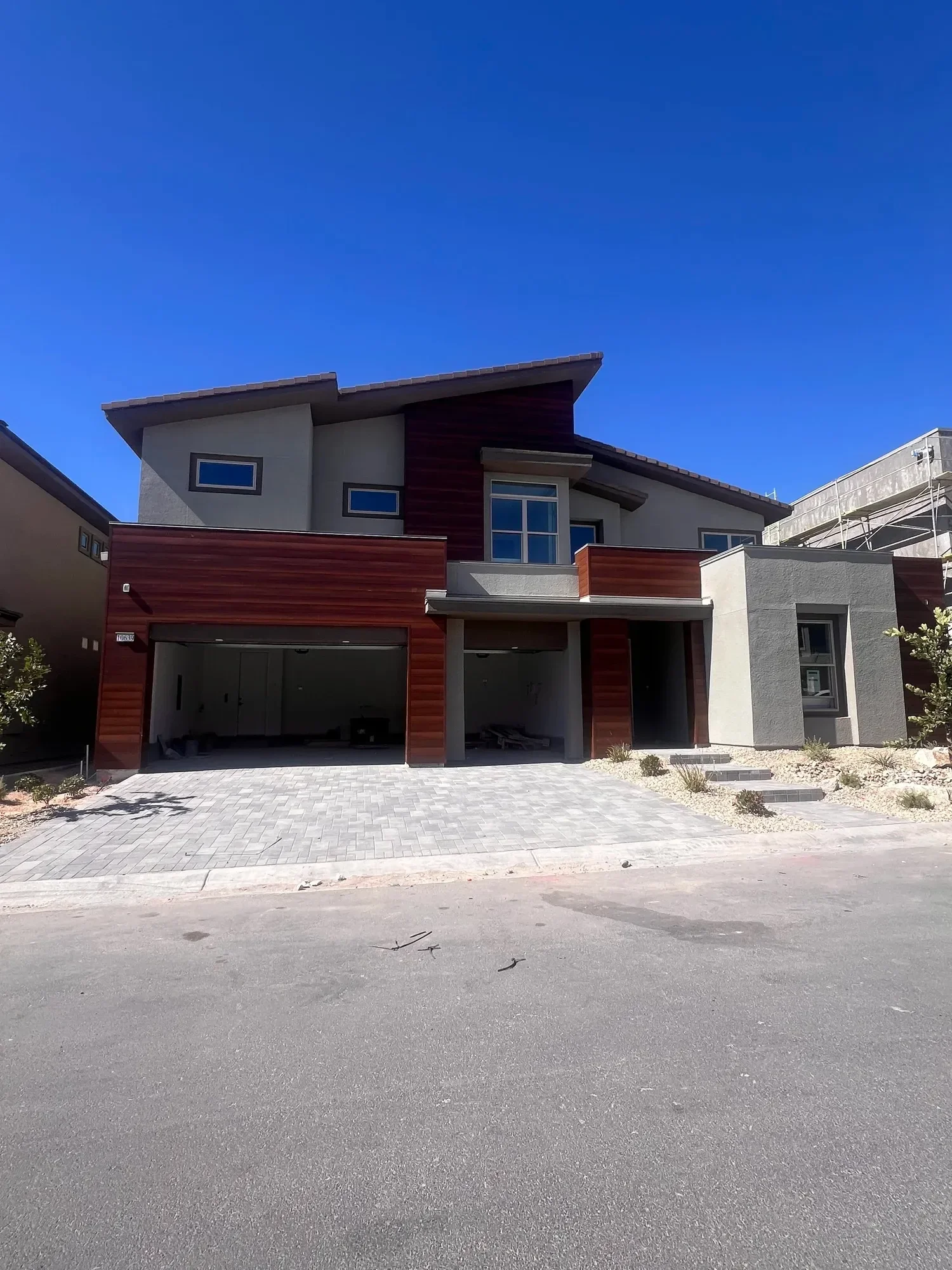 Modern two-story house with a gray and wood exterior, large windows, a garage, and a paved driveway under a clear blue sky.