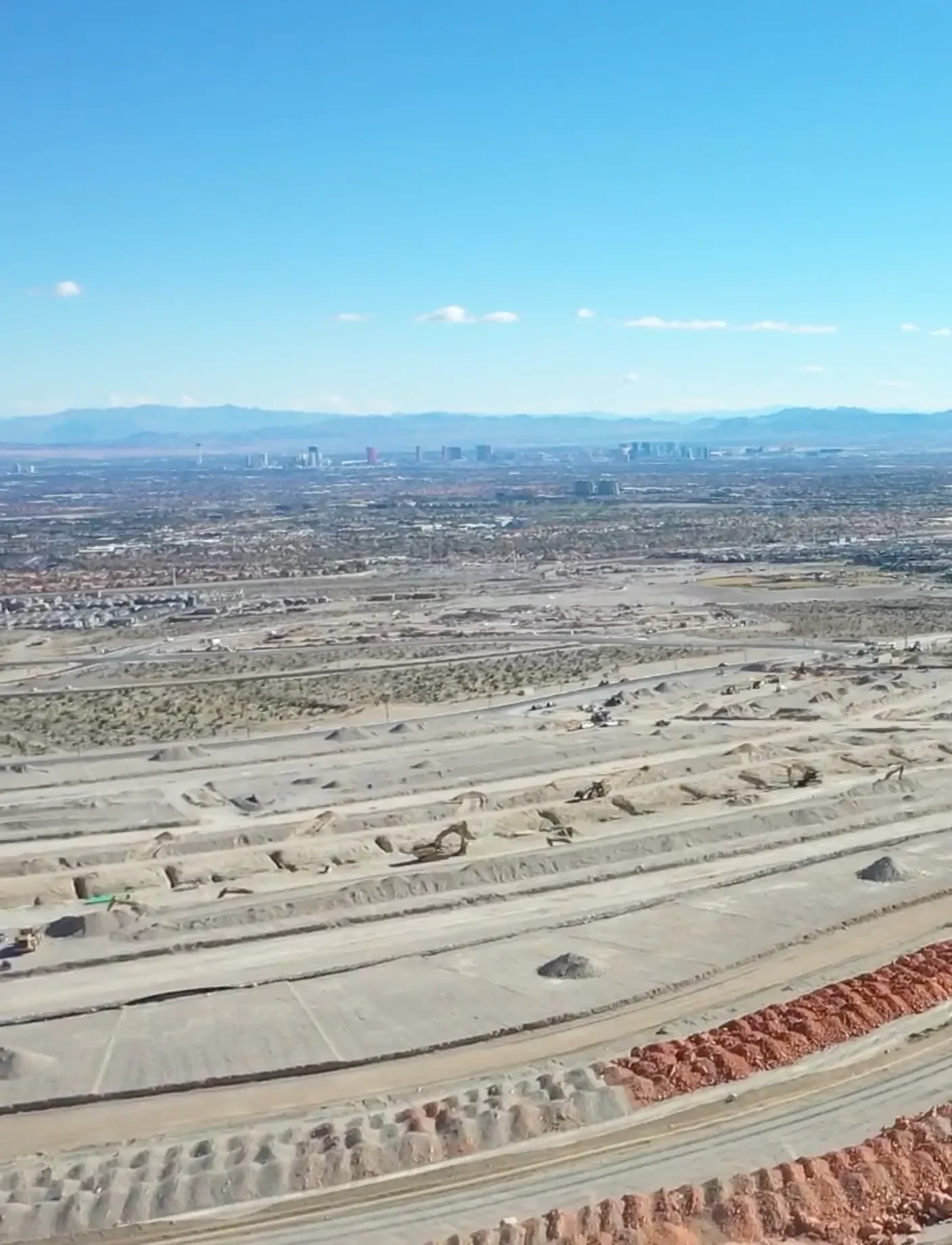 View of a city skyline from a high vantage point, with desert terrain and construction sites in the foreground, and mountains in the background under a clear blue sky.
