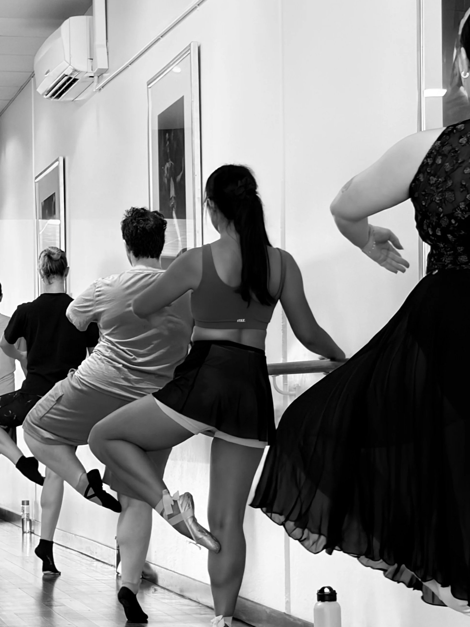Ballet dancers practicing at a barre in a dance studio with framed pictures on the wall, some dancers stretching or warming up, and water bottles on the floor.