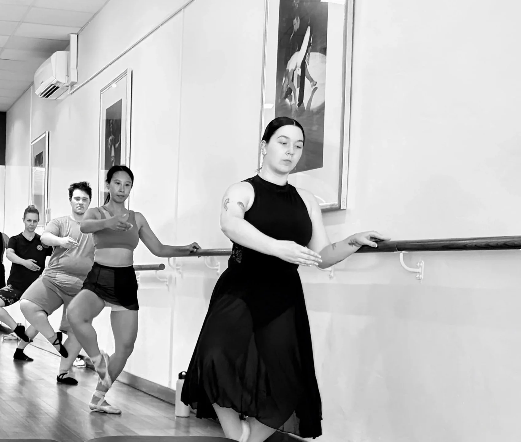 A group of five women practicing ballet at a studio, holding a ballet barre and focusing on their movements.
