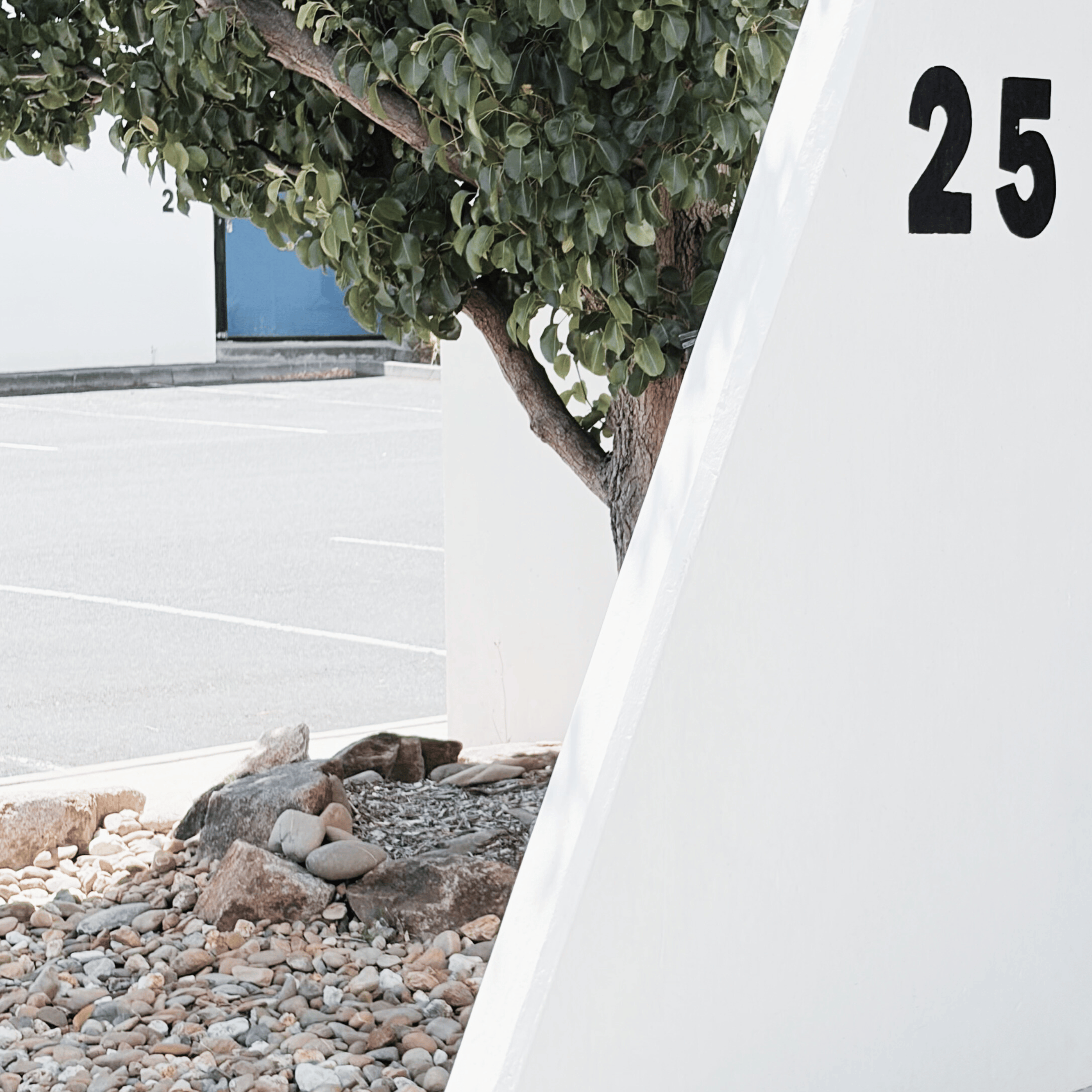 Partial view of a white building with the number 25, a tree with green leaves, rocks, and a parking lot in the background.