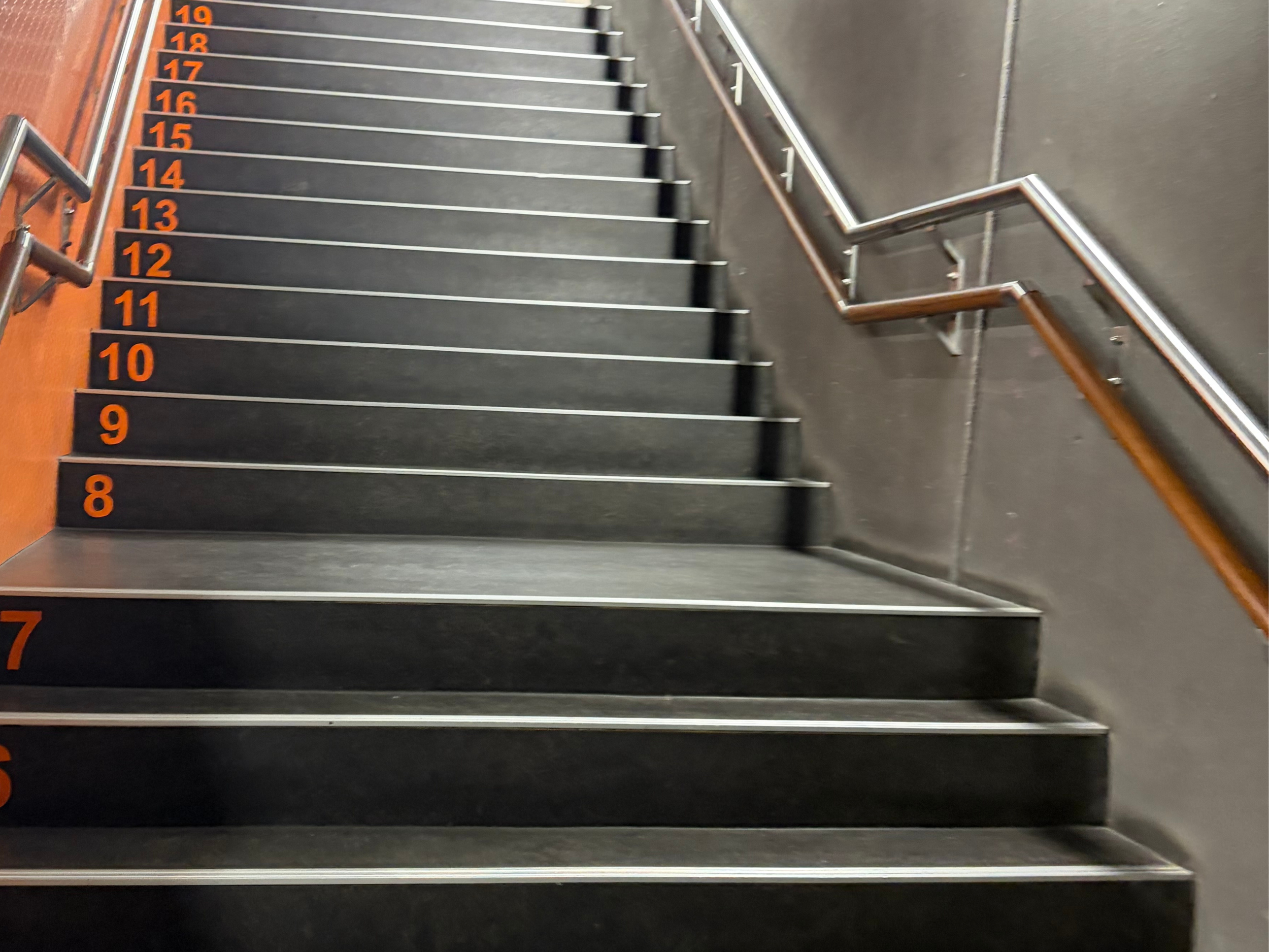Indoor staircase with black steps and orange numbering from 8 to 19 on the left side, with gray and wooden handrails on the right side.