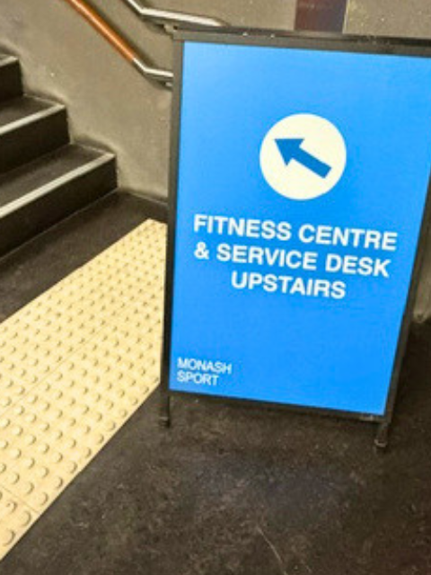 Blue signboard indicating direction to fitness center and service desk upstairs, near stairs with a tactile paving strip.