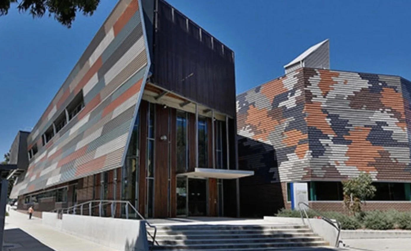 Modern building with angular design, featuring colorful patterned metal panels and large glass windows, with a staircase leading to the entrance, under a clear blue sky.