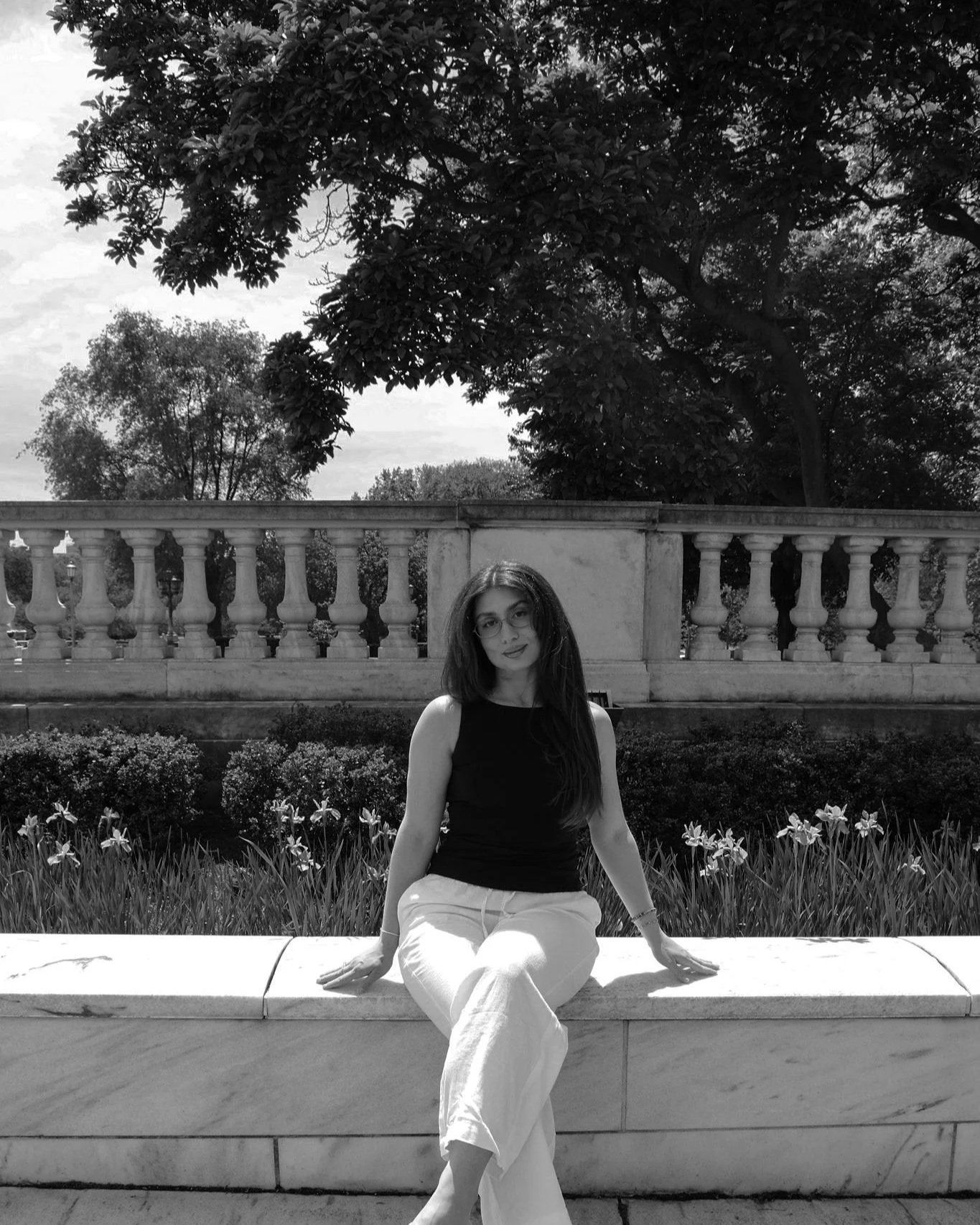 A young woman with long dark hair and glasses sitting on a stone bench outdoors. She is wearing a sleeveless black top and white pants, with her hands resting on the bench. Behind her, there is a decorative stone railing, lush grass, flowers, and trees under a cloudy sky.
