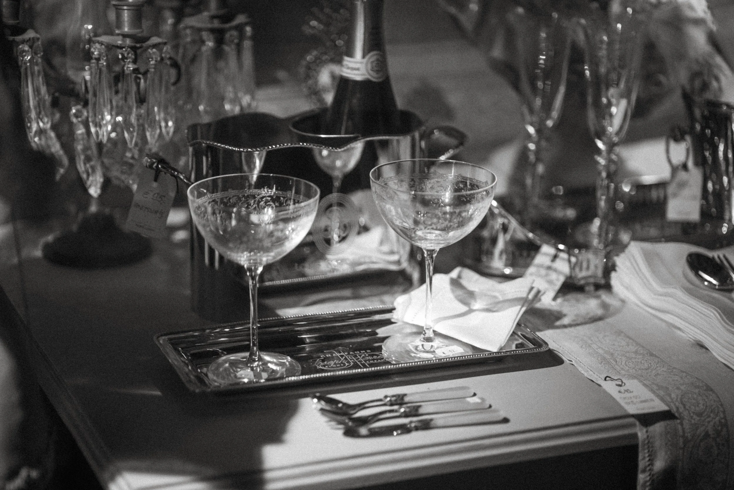 Black and white photo of a table setting with two empty cocktail glasses, silverware, and a tray, with various bottles and glasses in the background.
