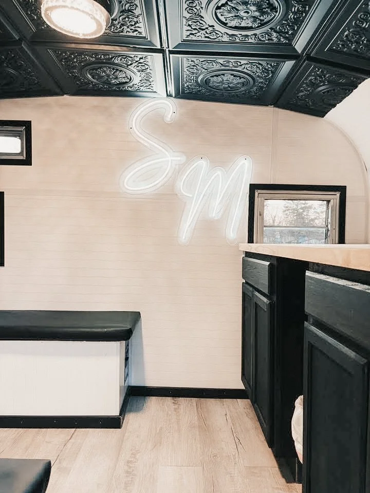 Interior of a room with beige walls, black cabinets, and a window. There is a neon sign on the wall that reads 'Spm' in cursive script, and decorative ceiling tiles.