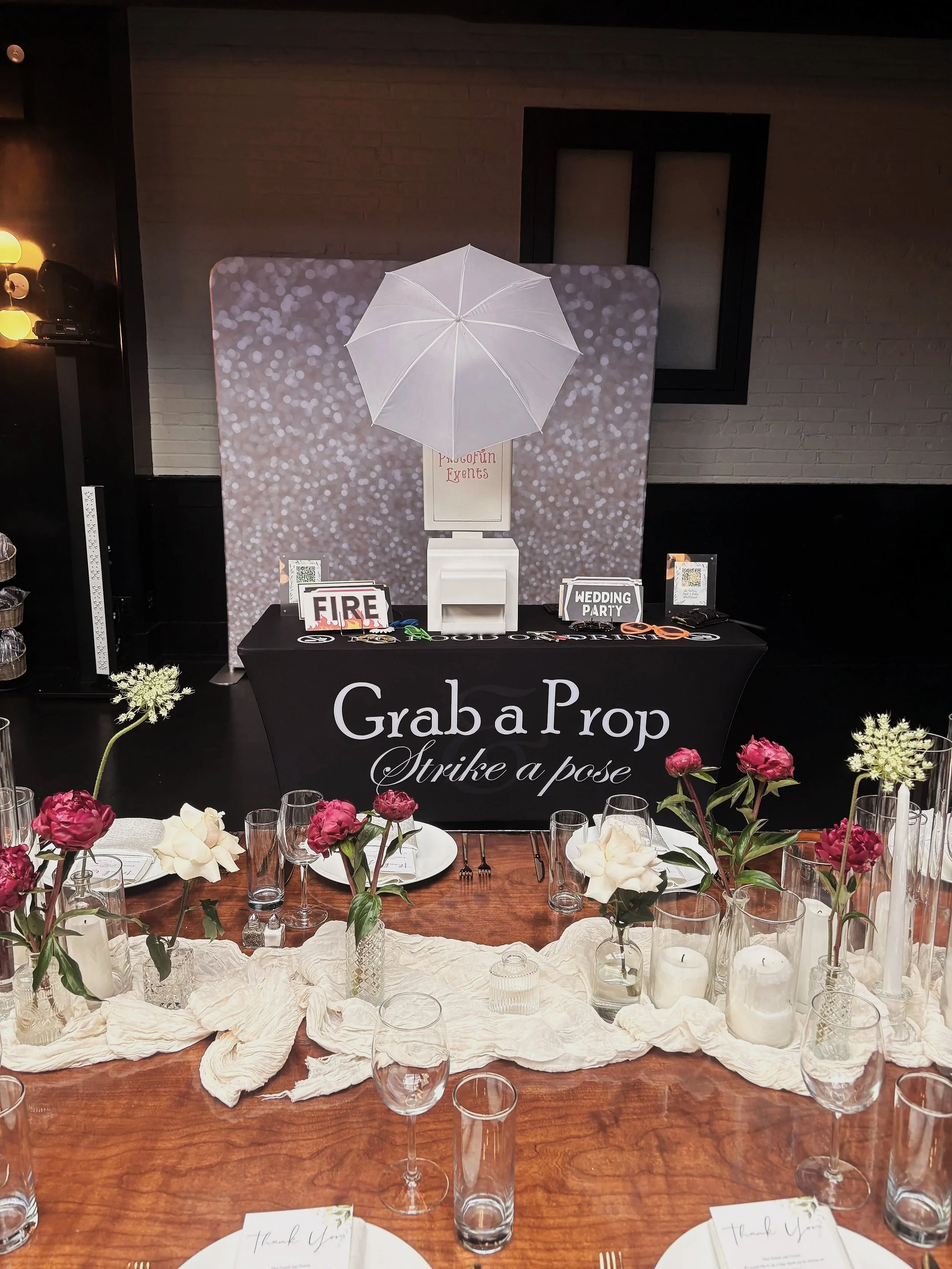 A reception table decorated with pink and white flowers in vases, votive candles, and glasses. There are place cards with 'Thank You' written. In the background, there is a photo booth with a sign that reads 'Photo Booth' and some props. The table has a black banner saying 'Grab a Prop, Strike a pose'.