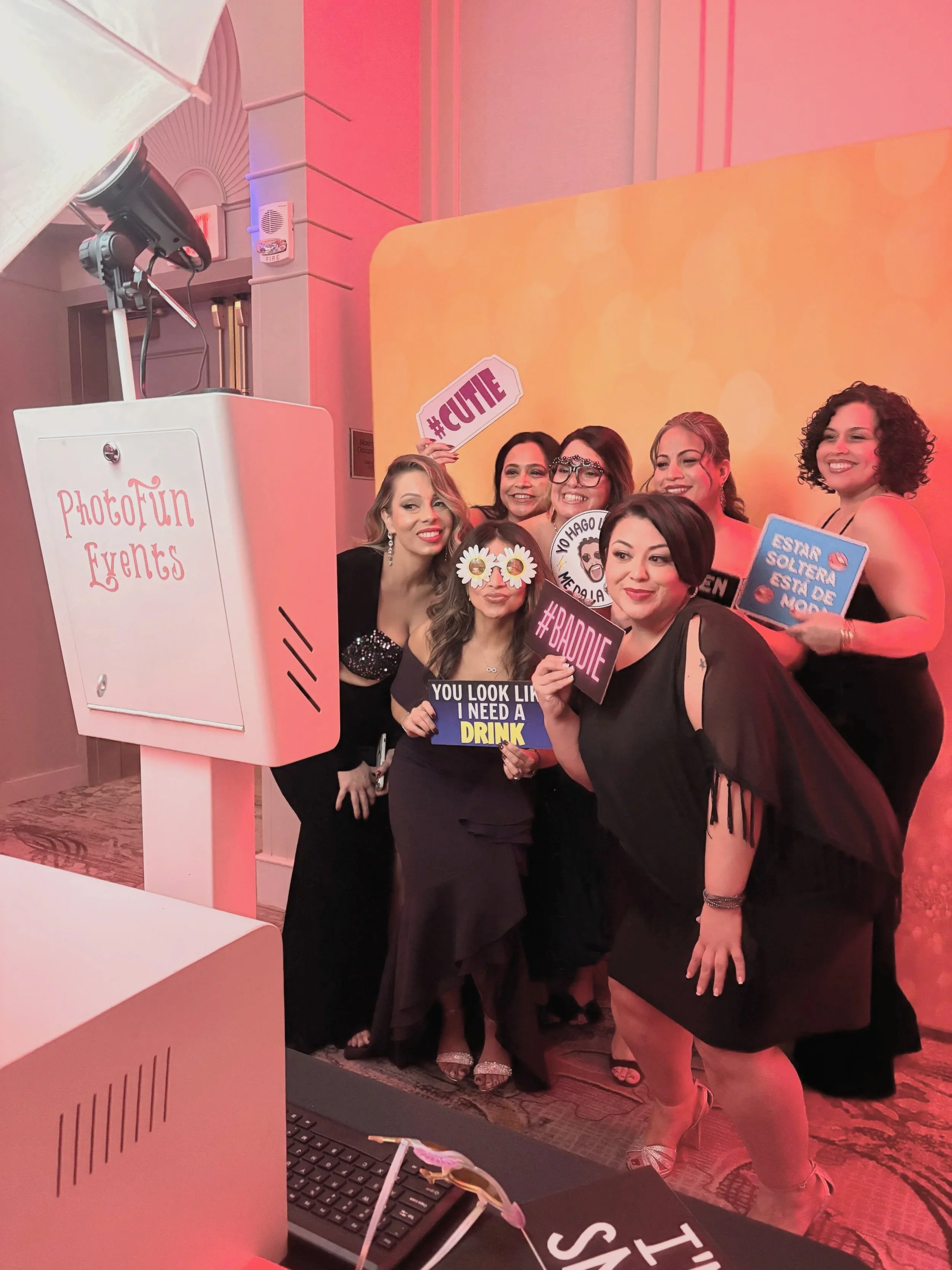 Group of women posing for a photo in a photo booth, holding signs with phrases like '#CUTE' and 'ESTÁ SOLTERA, ESTÁ DE MOOD.' They are dressed elegantly with some wearing glasses and accessories. The photo is taken indoors with pink lighting, and a sign on a pink booth reads 'PhotoFun Events.'