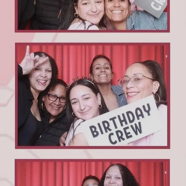 Group of five women smiling for a photo at a birthday celebration, with one holding a sign that says 'Birthday Crew', standing in front of a red curtain.