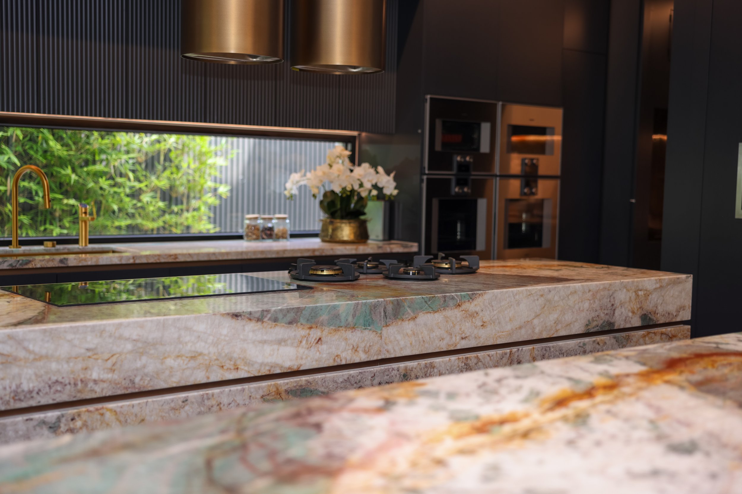 Modern kitchen with marble countertops, black stovetop, and gold fixtures, featuring a window with greenery, a vase of white flowers, and built-in oven and microwave.