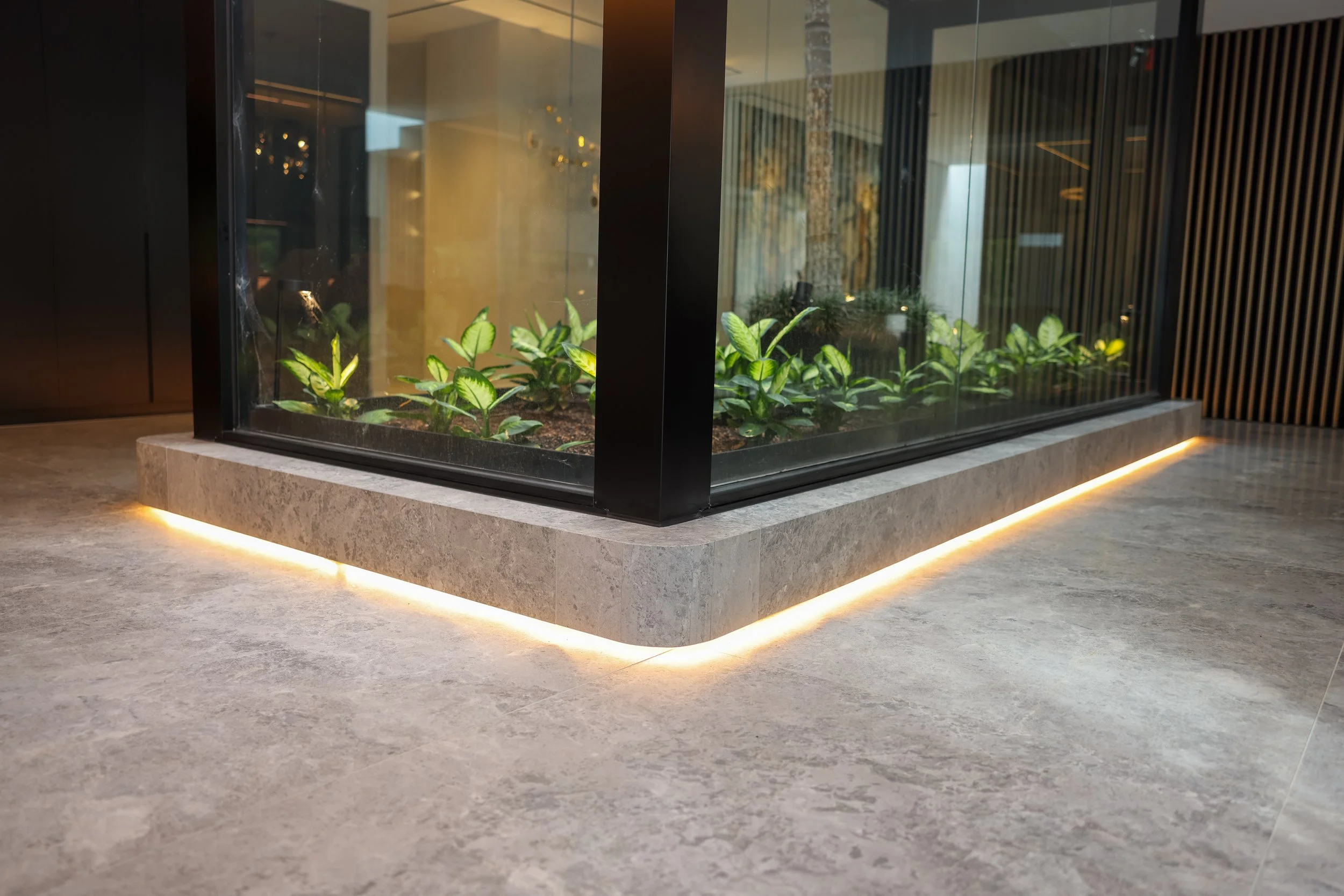 Indoor planter with green plants inside a glass enclosure, illuminated from below, in a modern interior with tiled floor and wooden wall panels.