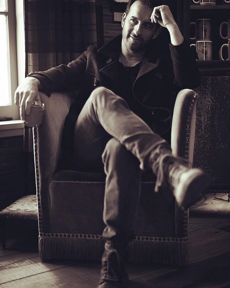 A man sitting in a cozy room by a window, smiling and resting his head on his hand. He has short dark hair and a beard, wearing a dark jacket and light-colored pants. He sits relaxed in an armchair with his legs crossed, holding a cup in his hand.