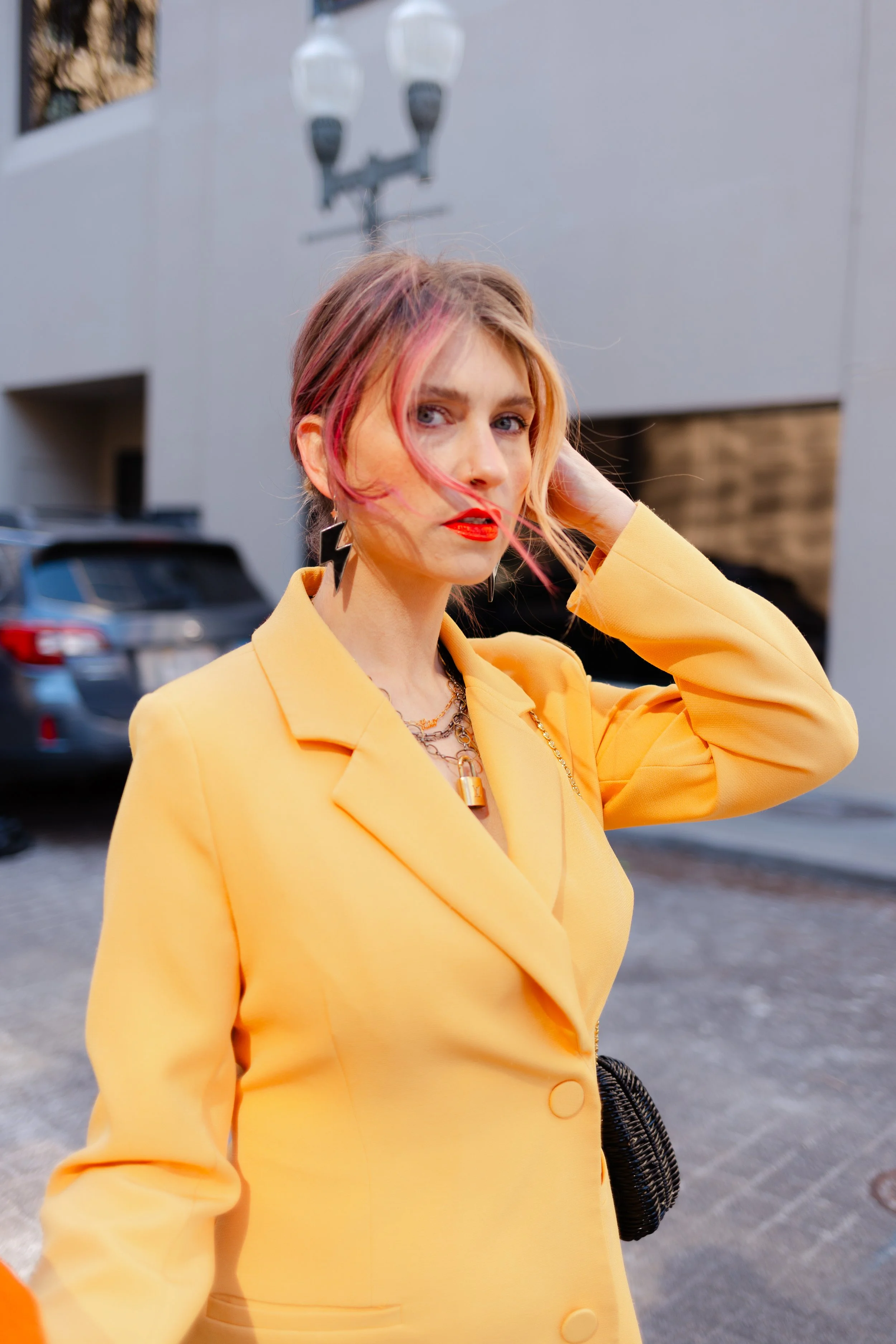 A woman with pink hair and red lipstick wearing a yellow blazer, standing outdoors with parked cars and a building in the background.