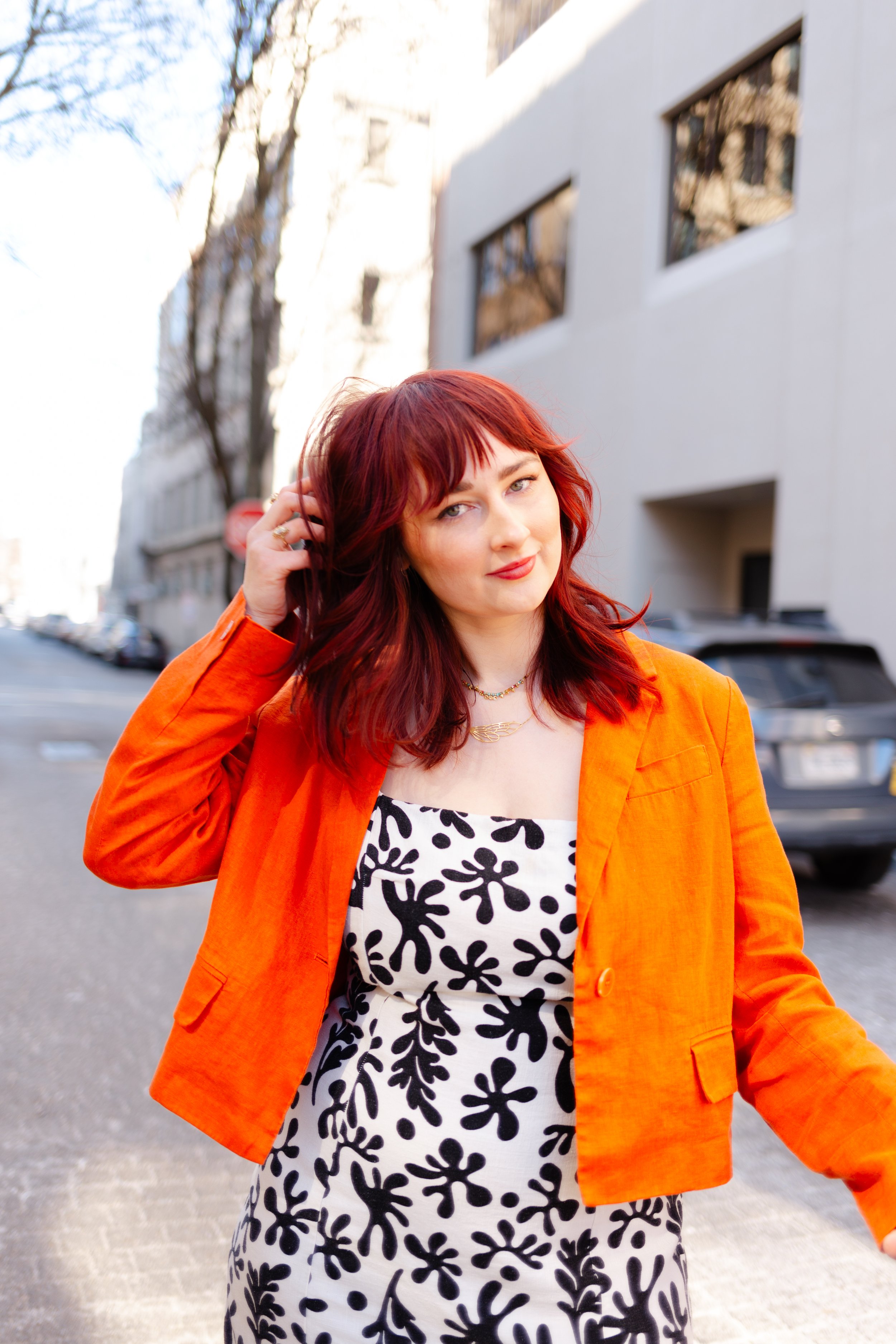 Woman with red hair wearing an orange jacket and black and white patterned dress standing on city street