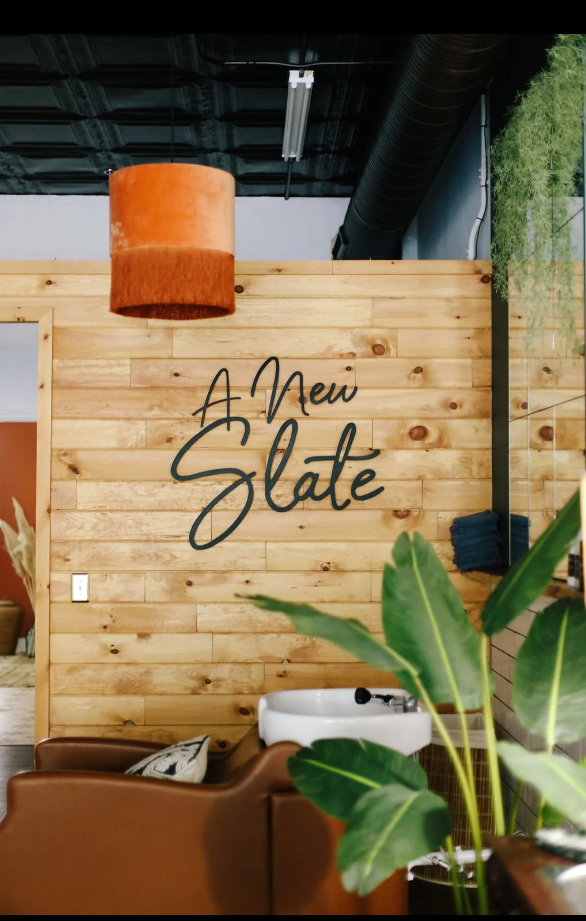 Interior of a salon or spa with a wooden wall, large green plant in the foreground, sign on wall reading 'A New Slate', ceiling with black ductwork and orange pendant light.