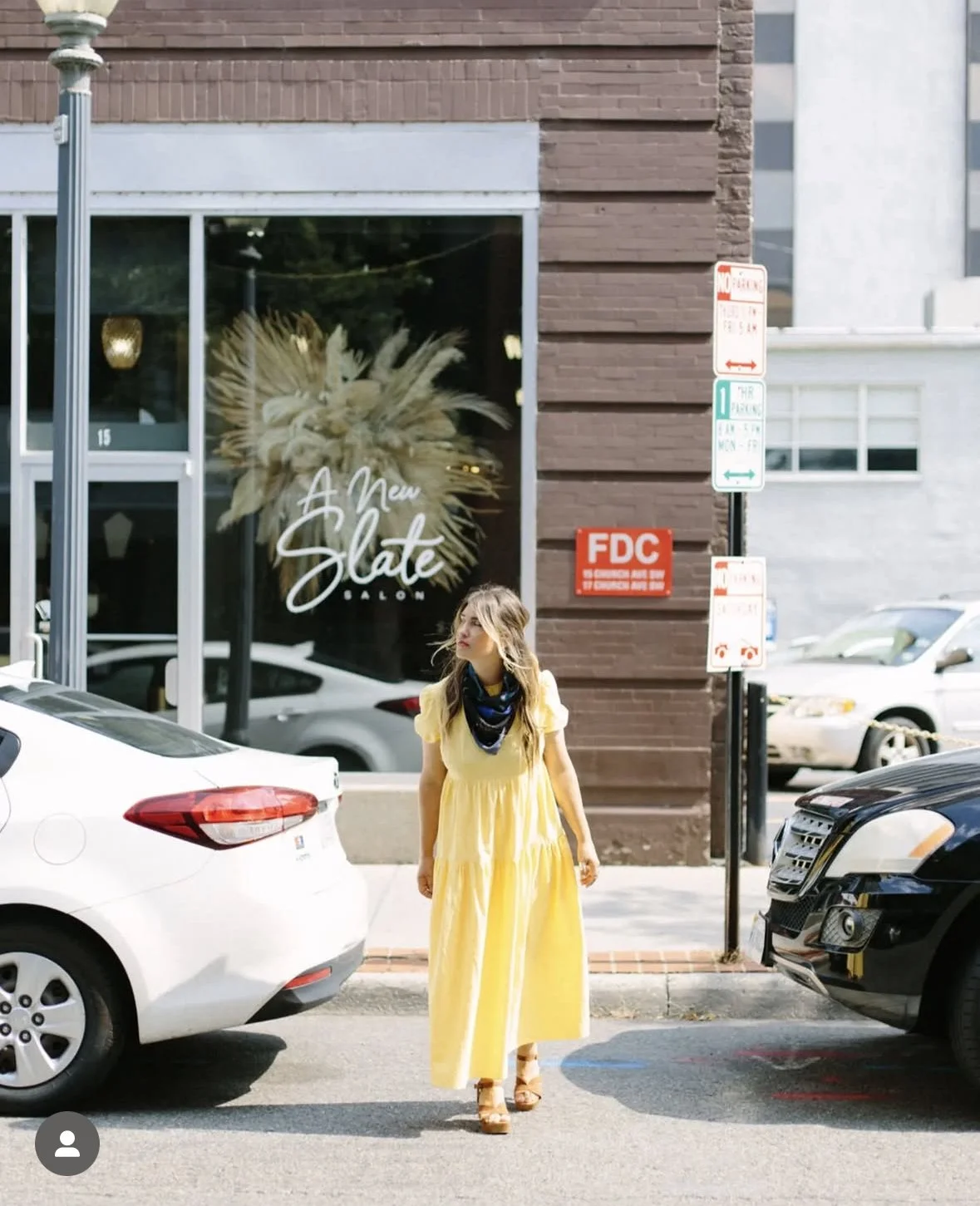 Woman in a yellow dress with a blue scarf walking past parked cars outside a salon called 'A New Slate Salon' in an urban setting.