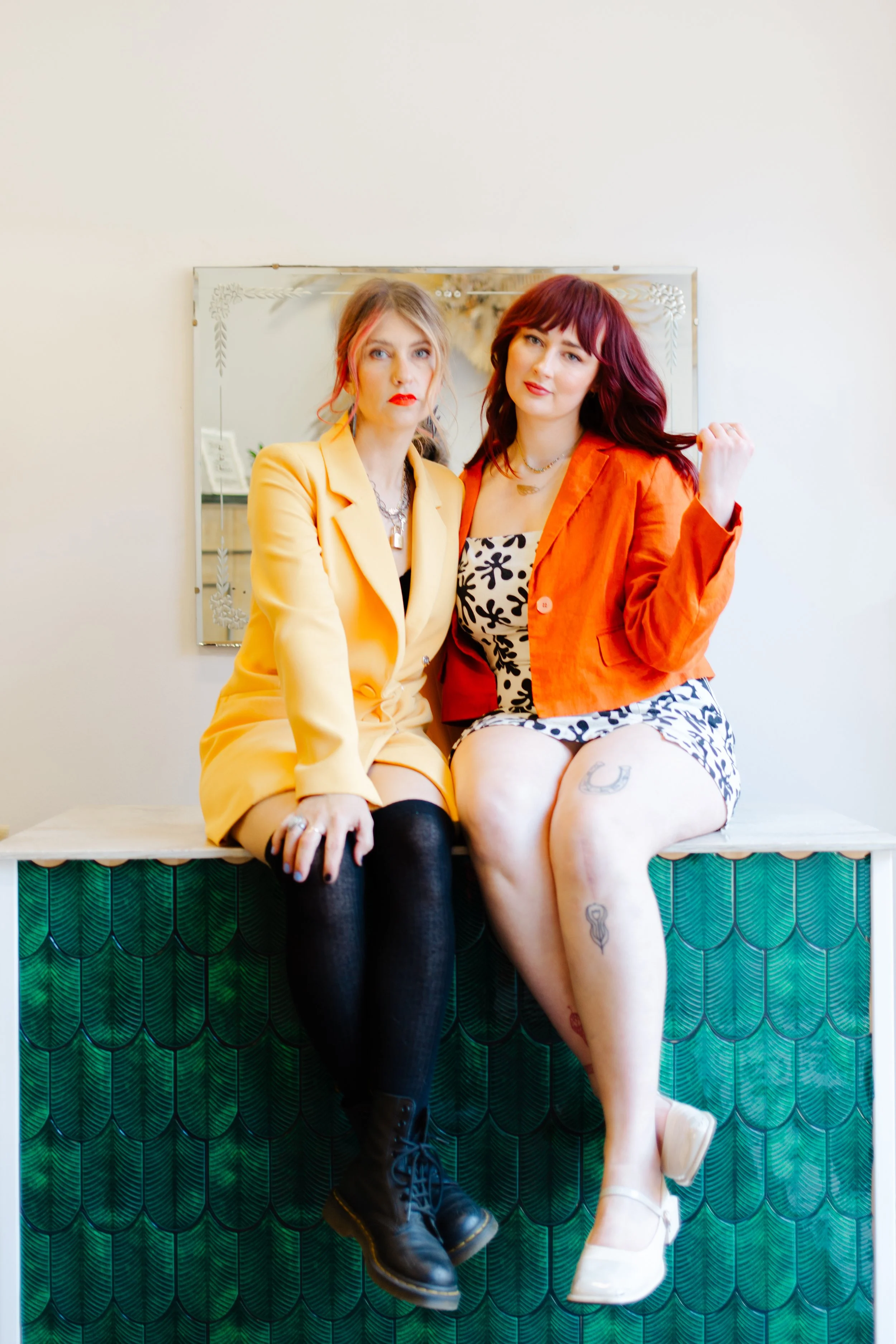 Two women sitting on a green patterned bench, posing for a photo indoors with a mirror behind them. One woman is wearing a yellow blazer and black boots, and the other is in an orange jacket and patterned dress, showing tattoos on her leg.