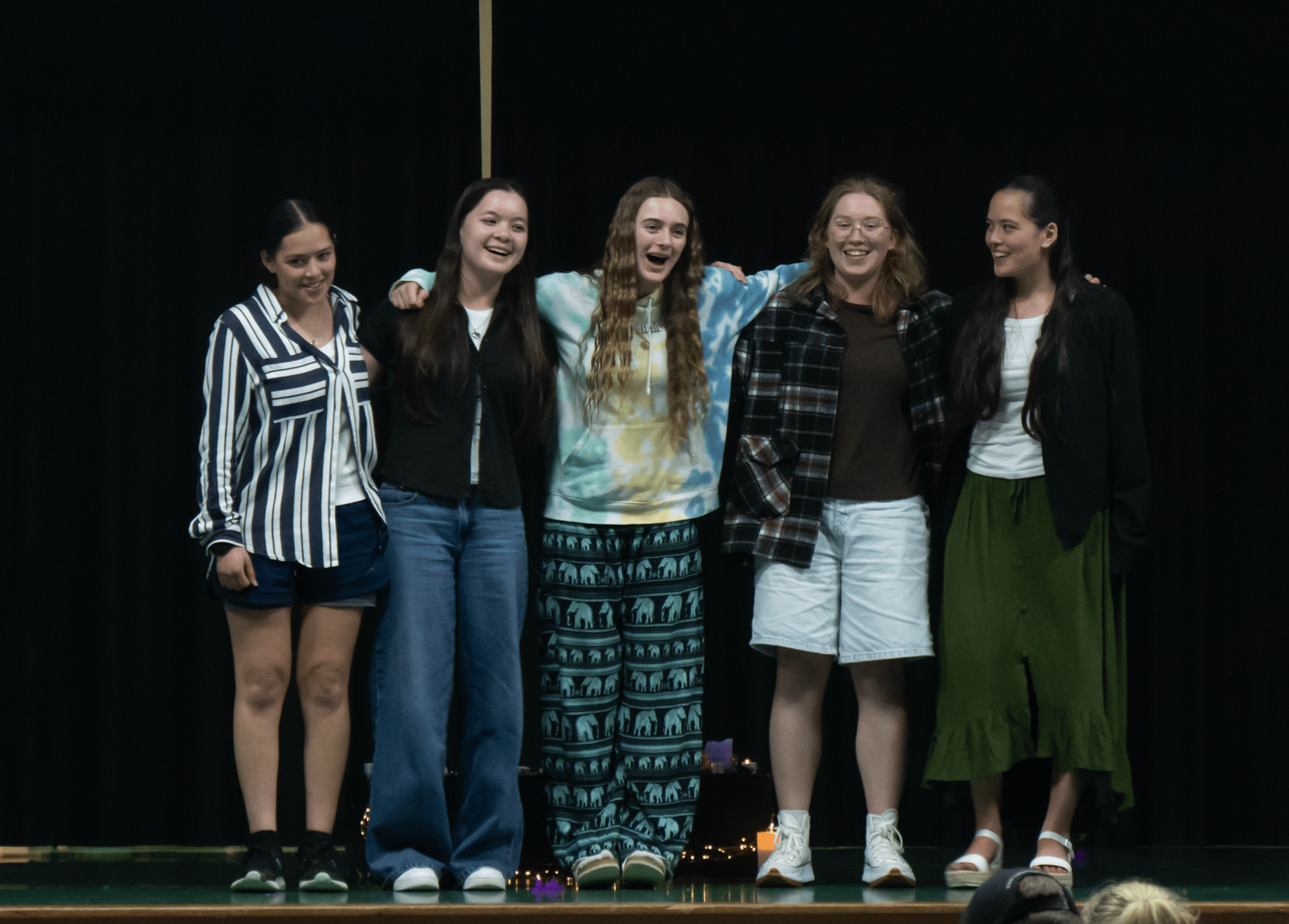 Five young women standing together on stage with arms around each other, smiling, in front of a black curtain.