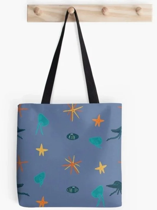 A tote bag with starfish, jellyfish, and kina sea urchin illustrations hanging from a hook on a wooden wall.